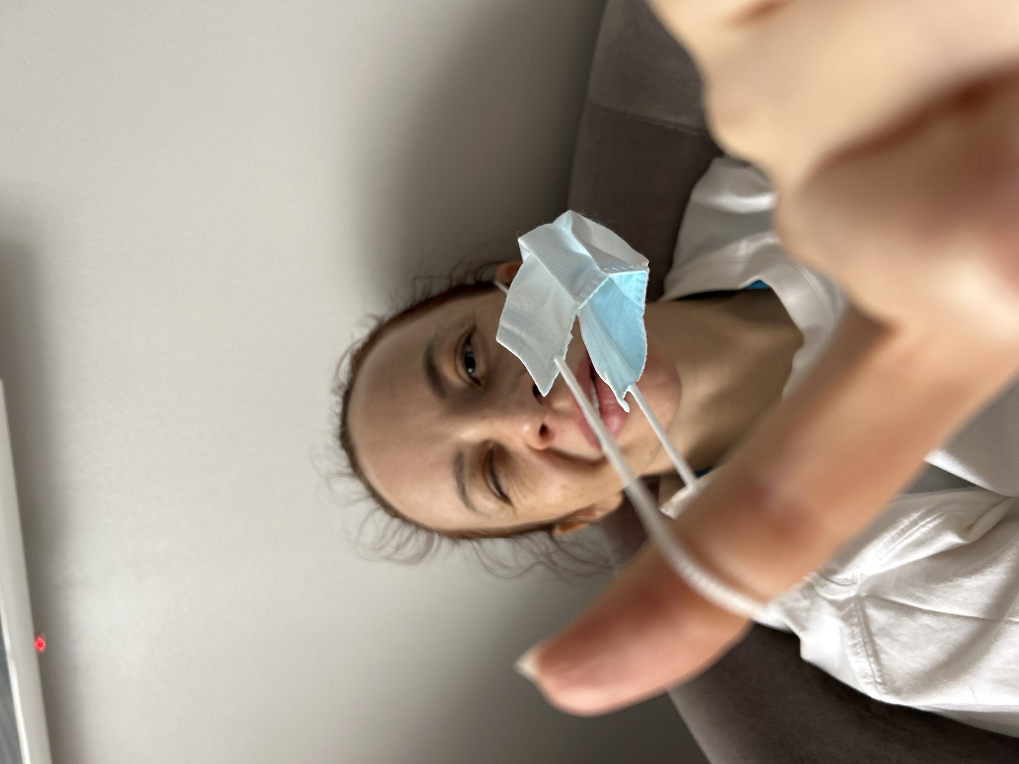 A woman is holding a surgical mask in front of her face. She is on a brown couch. The location of the city of Chaikovsky, Russia is not determined from the image.