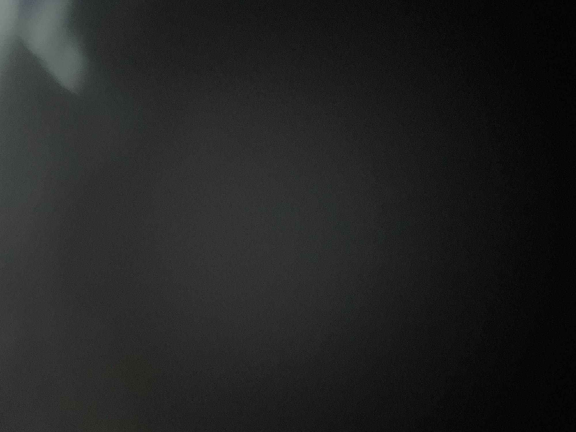 This is a still image, likely a photograph, with a dark, textured background. There are no discernible subjects such as people or objects. The lighting appears to be coming from the upper left, casting a bright, diffuse glare on that portion of the image. The rest of the image is predominantly dark, with subtle variations in tone that suggest a textured surface or grain. There is no visible text. The overall impression is abstract due to the lack of identifiable elements. The provided location context of Belgrade, Serbia does not offer any visual cues within the image itself.