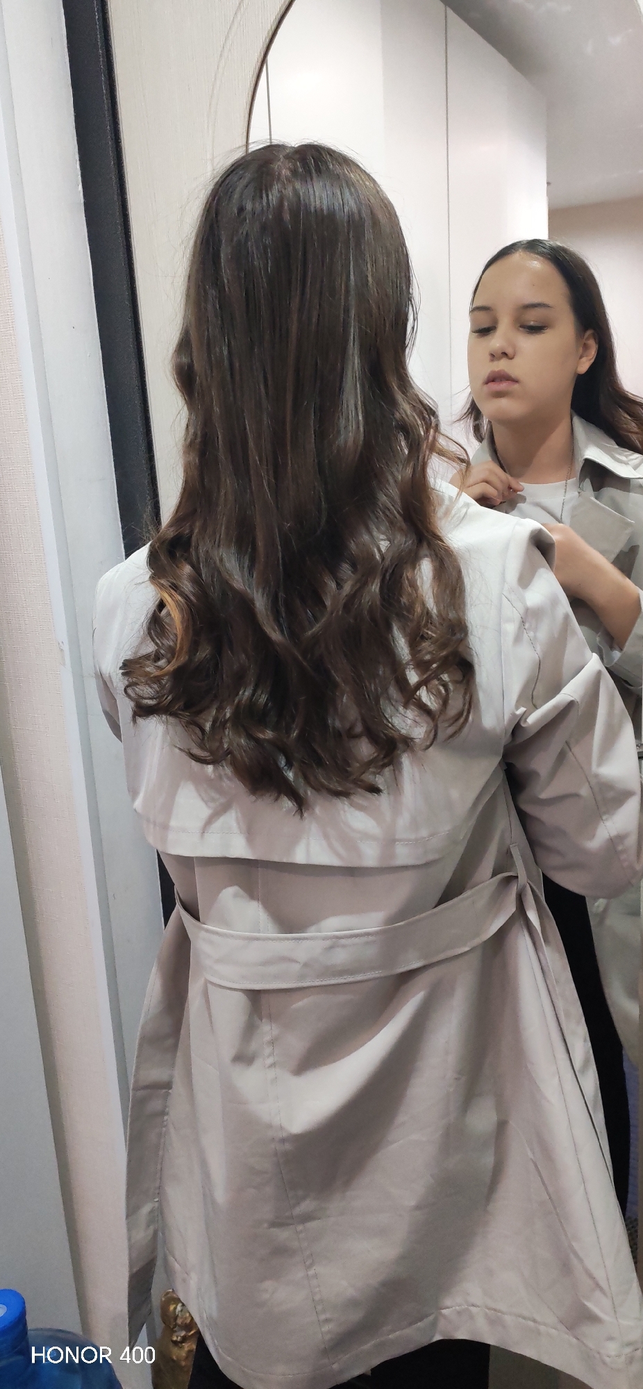 The scene is an indoor setting in Noviy, Russia. A female individual is positioned with her back partially towards the viewer, facing a large, dark-framed mirror. Her long, wavy, dark brown hair with visible lighter highlights extends down to her upper back. She is dressed in a light beige trench coat, which features a storm flap across the upper back and a belt tied loosely at the waist.
In her reflection within the mirror, the individual is seen adjusting an item around her neck, possibly a delicate chain necklace or the collar of an underlying light-colored (likely grey or white) top, using both hands. Her facial expression in the reflection is neutral with her eyes directed downward or forward, and her lips slightly parted.
The immediate environment visible includes light-colored walls; a section to the left of the individual has a subtle textured pattern, while the area reflected in the mirror appears to be a smooth, white wall or cabinet. In the lower left corner of the original image, a partial blue object, resembling a plastic bottle, and a blurry, dark object are visible. The watermark "HONOR 400" is also present in this corner. The lighting is uniform and consistent with an indoor environment.