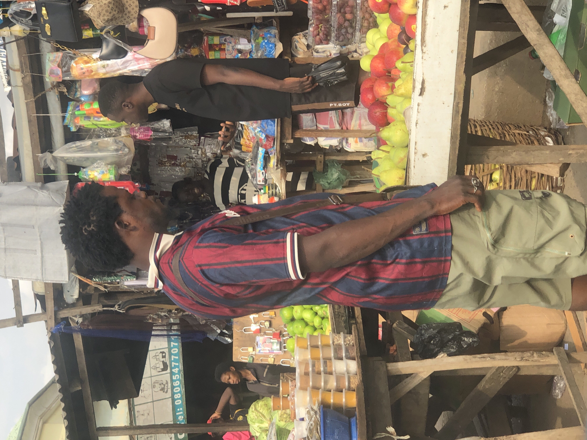In a bustling outdoor market stall in Lagos, Nigeria, two men stand engaged in conversation. The man in the foreground, with dark, curly hair, wears a red and blue striped short-sleeved shirt and light green cargo shorts. He has a dark strap across his chest and a ring on his right hand. The second man, facing away from the viewer, wears a black t-shirt and black shorts. He holds a black plastic bag.
The stall is laden with goods. To the left, stacks of clear plastic containers hold various food items, some appear to be spices or grains. A large basket filled with green vegetables, possibly cabbage, is visible behind the containers. Further back, a display of green mangoes or similar fruit sits on a wooden shelf alongside several padlocks.
To the right of the stall, a variety of fresh produce is displayed. Pears and apples, in shades of green, yellow, and red, are piled on a white surface. Behind the fruit, a stack of red grapes in plastic containers is visible.
The market environment is evident with the wooden structures of the stalls, overhead coverings, and various items hanging from the ceiling, including bags and what appear to be children's toys or novelty items. The ground surface seems to be a mixture of dirt and possibly pavement. The overall scene depicts a vibrant market setting with an emphasis on the commercial exchange of goods.