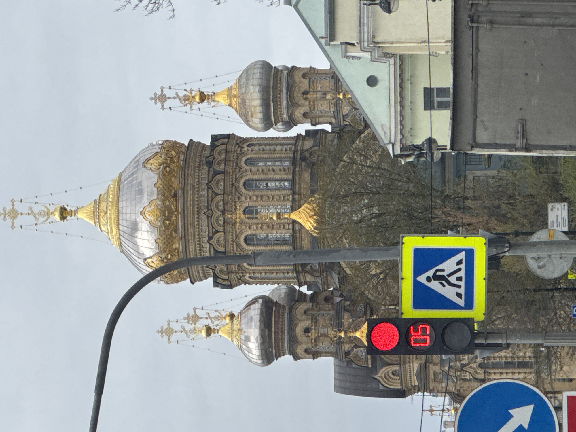 The image, rotated 90 degrees clockwise, captures a detailed urban scene in Saint Petersburg, Russia. The central subject is a magnificent Russian Orthodox cathedral, characterized by its multiple ornate golden and silver-grey onion domes, topped with intricate Orthodox crosses. The cathedral's facade features elaborate architectural detailing, including arched windows and decorative patterns.
In the foreground, a prominent street pole holds several traffic-related objects. A traffic light displays a red signal alongside a digital countdown showing "50," indicating the remaining time for the red light. Above the traffic light is a yellow-bordered blue square sign depicting a white triangle with a black pedestrian icon, signifying a pedestrian crossing. Below the traffic light, a circular blue sign with a white arrow pointing right is partially visible.
The background consists of a uniform, overcast grey sky. Bare tree branches, suggesting late autumn or winter, are visible to the right of the cathedral. Portions of other buildings, including a lighter-colored structure with windows and a darker, more industrial-looking building, are seen on the right side of the frame. No people are discernible in the image. The scene conveys a typical daytime urban environment, highlighting the distinctive Russian Orthodox architecture amidst city infrastructure.