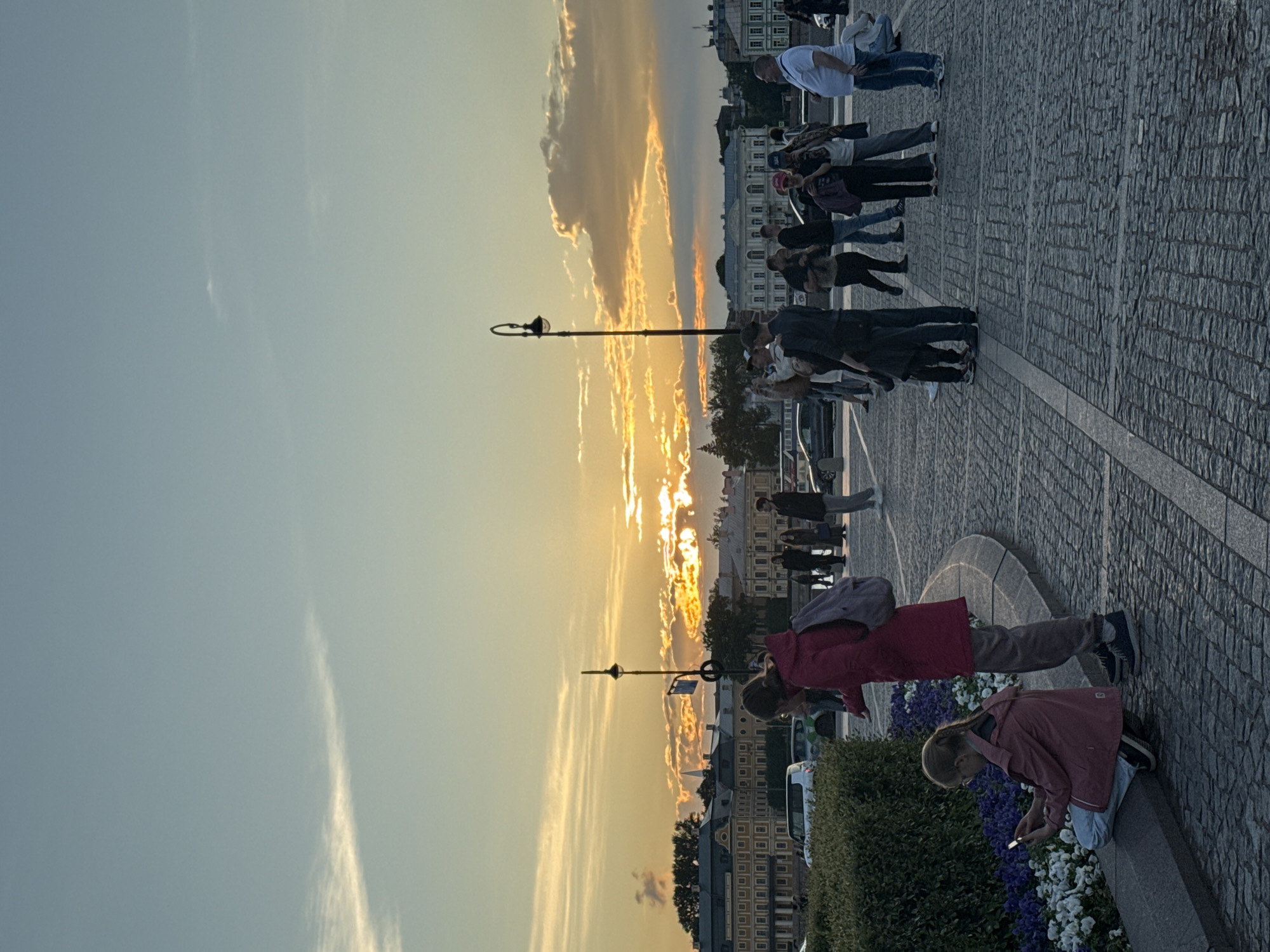 A crowd of people stand on a cobblestone promenade in Saint Petersburg, Russia, during sunset. The sky is a mix of blue and orange hues, with dramatic clouds illuminated by the setting sun. In the background, ornate, historic buildings line the horizon, characteristic of the city's architecture. Several individuals are taking photos or looking at their phones, while others are engaged in conversation. A lamppost stands tall on the promenade, and a woman in a red coat with a backpack stands near a flowerbed where a young girl, also in a pink jacket, is sitting and looking at her phone. Cars and trees are visible in the distance. The scene captures a moment of leisure and sightseeing in a picturesque urban setting.