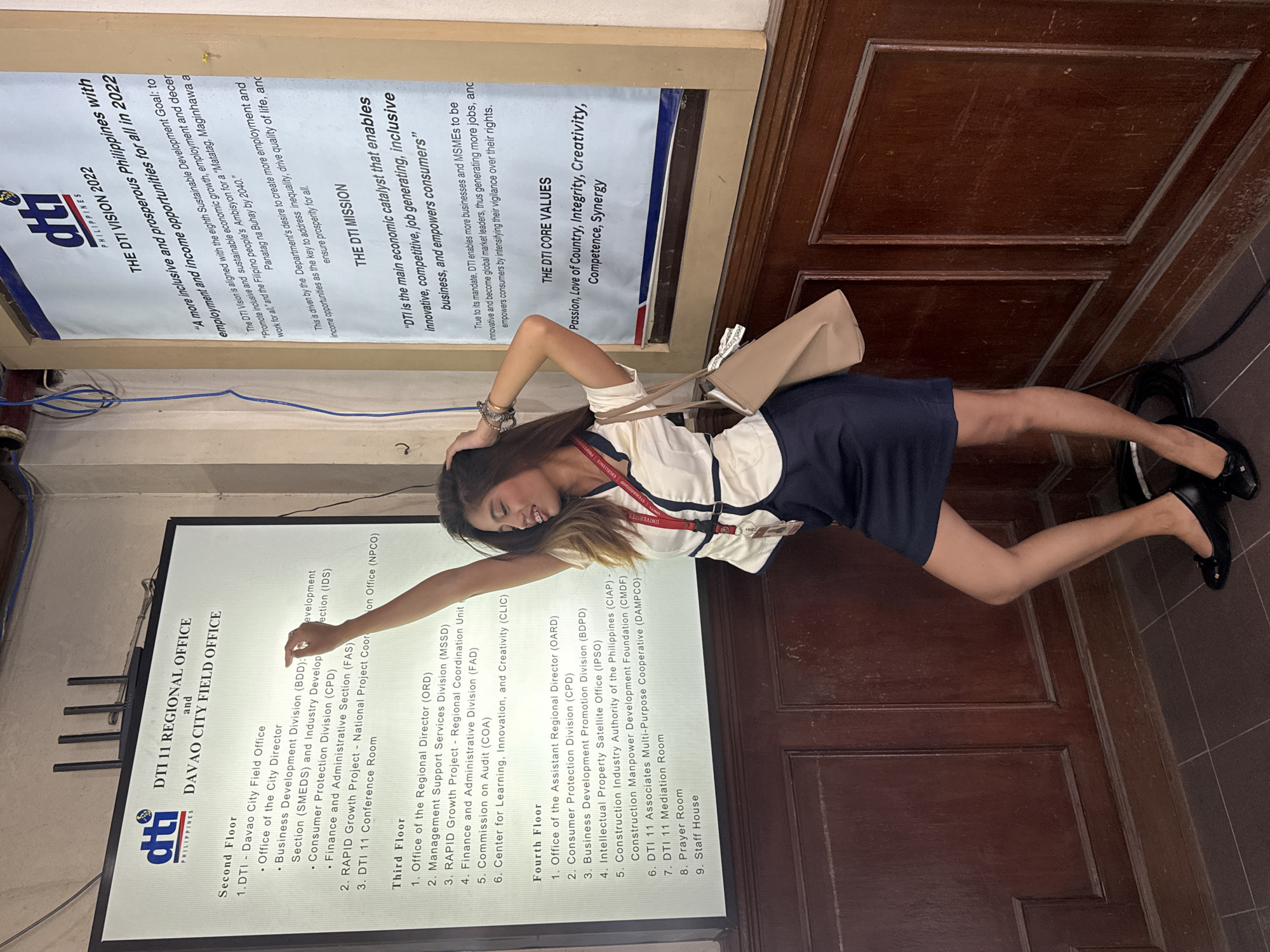 The image depicts a young woman standing in an indoor space, possibly an office or government building, in Davao City, Philippines. She is positioned in front of a large, illuminated signboard displaying organizational information for "DTI 11 REGIONAL OFFICE and DAVAO CITY FIELD OFFICE". The signboard details office locations by floor, including the Second, Third, and Fourth Floors, with specific divisions and departments listed.
The woman is wearing a white collared shirt with dark trim and a dark skirt, and she has a lanyard around her neck with an ID card. She is holding a light-colored handbag. Her posture suggests she is posing for a photograph, with one arm raised and her head tilted back.
To the right of the signboard, a large banner is partially visible, featuring text related to "THE DTI VISION 2022" and "THE DTI MISSION," along with "THE DTI CORE VALUES."
The environment appears to be well-lit, with wooden paneling on the walls below the signboards. The floor is tiled. The overall impression is that of an official government office setting within Davao City.