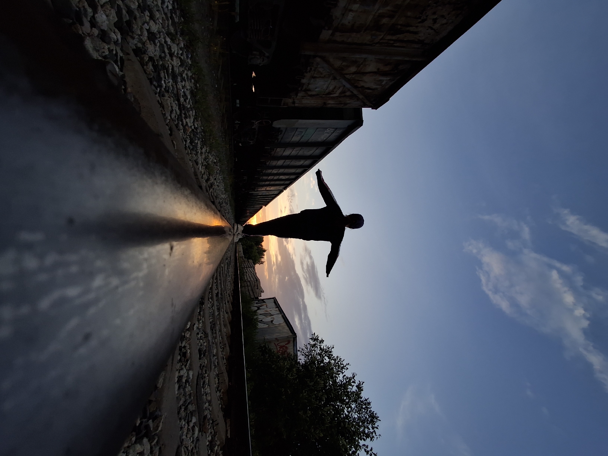 A person silhouetted against a sunset walks along a railway track. The person, seen from behind, has their arms outstretched to maintain balance. They are wearing dark clothing and white sneakers. In the background, to the left, are several dark freight train cars. To the right, there are trees and a corrugated metal structure covered in graffiti. The sky is a gradient of blue and orange, with some wispy clouds. The foreground is dominated by a close-up of the shiny metal railway track, reflecting the sky and sunset. The surrounding area consists of gravel ballast. The scene appears to be located in an industrial or rail yard area. Given the context of Chaikovsky, Russia, this could be a railyard on the outskirts of the city.