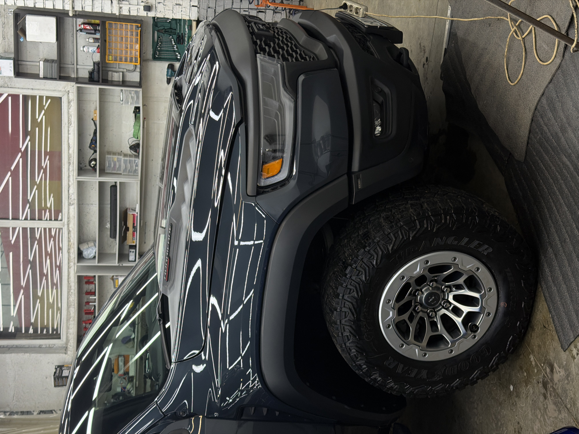 The image displays the front-right section of a dark grey/blue Ram TRX pickup truck, parked indoors within what appears to be a professional garage or detailing facility. The vehicle's highly polished surface vividly reflects linear overhead lighting. Visible features include a prominent hood scoop, a "TRX" decal on the hood's power bulge, an aggressive black grille, LED headlights with amber turn signals, black fender flares, and a multi-spoke silver and black wheel fitted with a Goodyear Wrangler off-road tire.
The environment consists of light-colored walls, with a window in the background reflecting the same linear ceiling lights. Various shelving units are present, holding an assortment of workshop tools, bottles, containers, and a yellow multi-compartment organizer. A green toolbox is situated beneath one shelf. A yellow electrical extension cord is coiled on the concrete floor, connecting to a white three-outlet power strip positioned near the truck's front bumper. The concrete floor exhibits some moisture or liquid beneath the vehicle. No individuals are visible in the scene. The specific location within Krasnoyarsk, Russia, is not identifiable from the visual information provided.