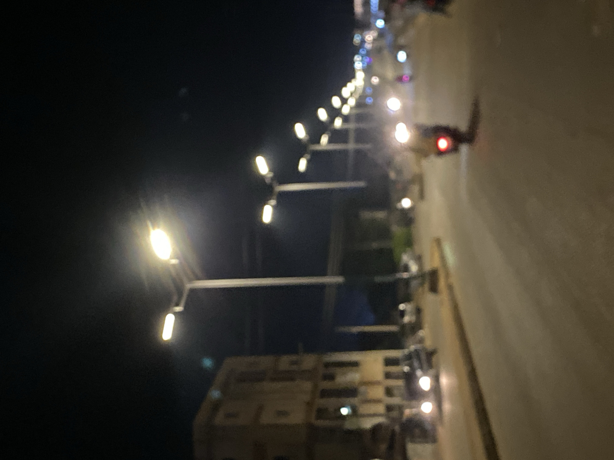 The image depicts a night scene on a street in Bukuru, Nigeria, illuminated primarily by a long row of streetlights. The perspective is tilted, showing the street stretching into the distance.
Numerous streetlights with bright, warm-toned illumination are prominent, lining what appears to be a paved road. These lights create a strong visual line, guiding the eye down the street. Further down the road, additional lights, some appearing cooler-toned and others as blurred points of light (bokeh), suggest more distant illumination or decorative lights.
On the right side of the image (if viewed upright), a portion of the road is visible, along with blurred shapes that appear to be buildings or structures adjacent to the street. A vehicle, likely a motorcycle, is visible in the mid-ground on the right, distinguished by its bright red taillight. Other blurred lights suggest additional vehicles or parked motorcycles/cars along the street. The sky above is dark, confirming it is nighttime. The overall scene is somewhat out of focus, giving a soft, dreamy quality to the lights. There is no visible text in the image.
