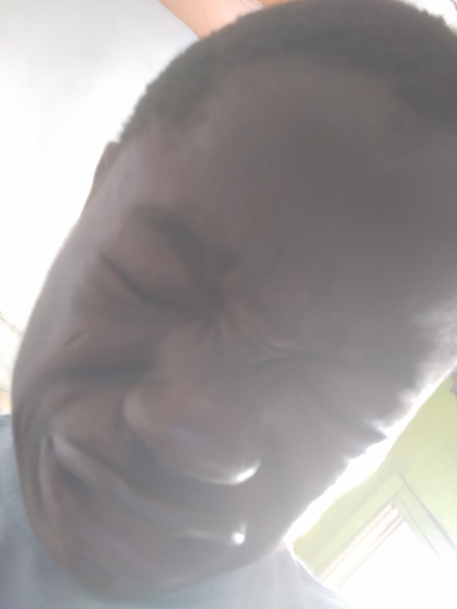 A close-up, low-angle photograph depicts a person with dark skin, seemingly squinting or closing their eyes. The lighting is bright and partially obscures the subject's features, particularly around the nose and forehead. A small portion of the subject's clothing is visible at the bottom of the frame. The background is out of focus, but a section of a light-colored wall and window frame are visible, suggesting an indoor environment. The exact location in Shangisha, Nigeria, cannot be determined from this image.