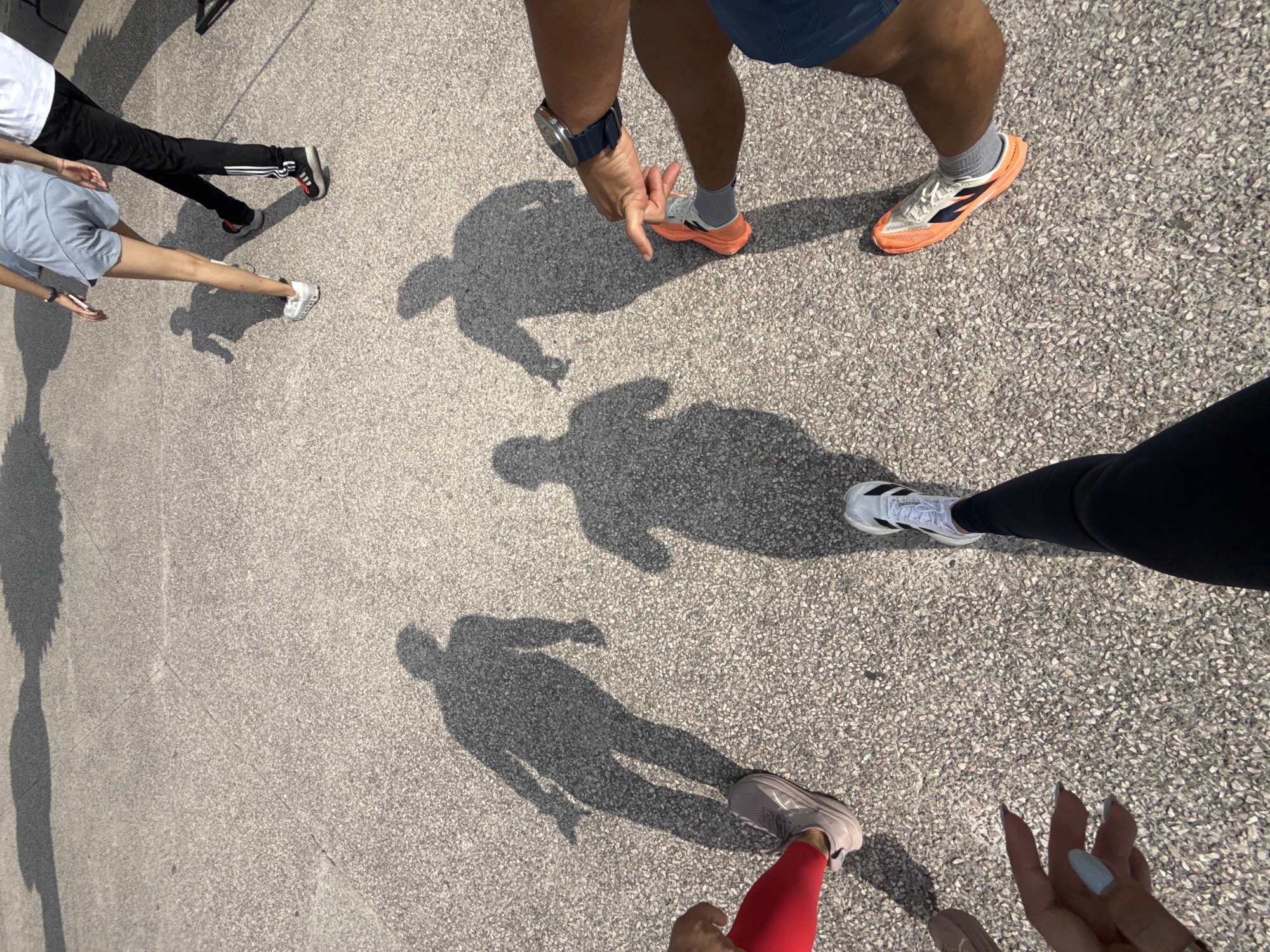 The image, likely taken in Puebla, Mexico, shows a section of textured grey pavement under bright sunlight, casting dark shadows of several people, presumably pedestrians or runners. The shadows indicate the presence of at least five individuals; however, only their lower body parts are visible in the frame. The person closest to the camera is wearing red leggings and light grey sneakers. To the right, an individual is wearing black leggings and white sneakers, with manicured fingers. Further in the background, there's a person in light blue shorts and white sneakers, and another in black athletic pants with Adidas sneakers. The top of the frame shows a person in blue shorts, grey socks, and orange-accented sneakers, their hand extended downward with a watch on their wrist. There's no explicit information confirming the image location to be Puebla, Mexico. It is possible that the image was taken in a different location.