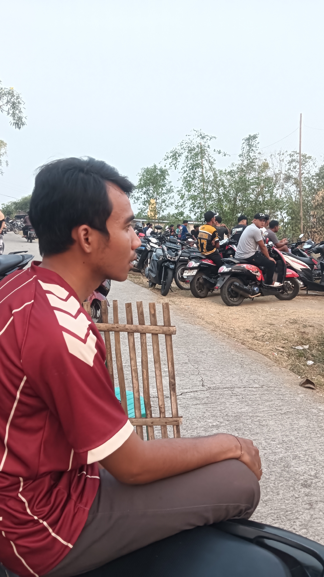 In Tanggan, Indonesia, a man with short black hair, wearing a maroon shirt with white chevron patterns and dark brown pants, is seated in the foreground, facing right. His right arm rests on his bent right leg, and his left arm is out of frame. Behind him, a group of people on motorcycles are parked along a road. The background features trees and a hazy sky. A low bamboo fence is visible to the left of the seated man.