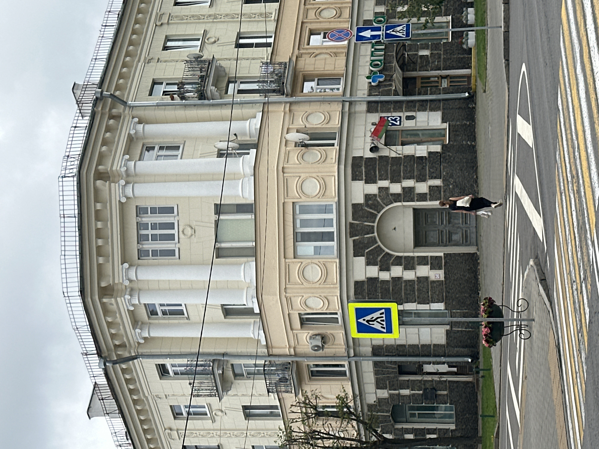 The image depicts a street scene in Minsk, Belarus, focusing on the exterior of a building with classical architectural elements, including columns and decorative moldings. The building is light yellow on the upper floors and dark stone on the lower facade. It has multiple windows, balconies with wrought-iron railings, and satellite dishes attached to the exterior. A pharmacy sign with Cyrillic lettering is visible on the right side of the building.
In the foreground, a pedestrian crossing is marked by white and yellow stripes on the asphalt. A woman dressed in black, carrying white shopping bags, is walking across the street on the pedestrian crossing, moving from left to right. A street sign indicating a pedestrian crossing is present to the left of the woman. On the sidewalk, a decorative planter with pink and dark red flowers sits near the pedestrian crossing. The sky is overcast and gray. The image is taken from a low angle, looking up towards the building.