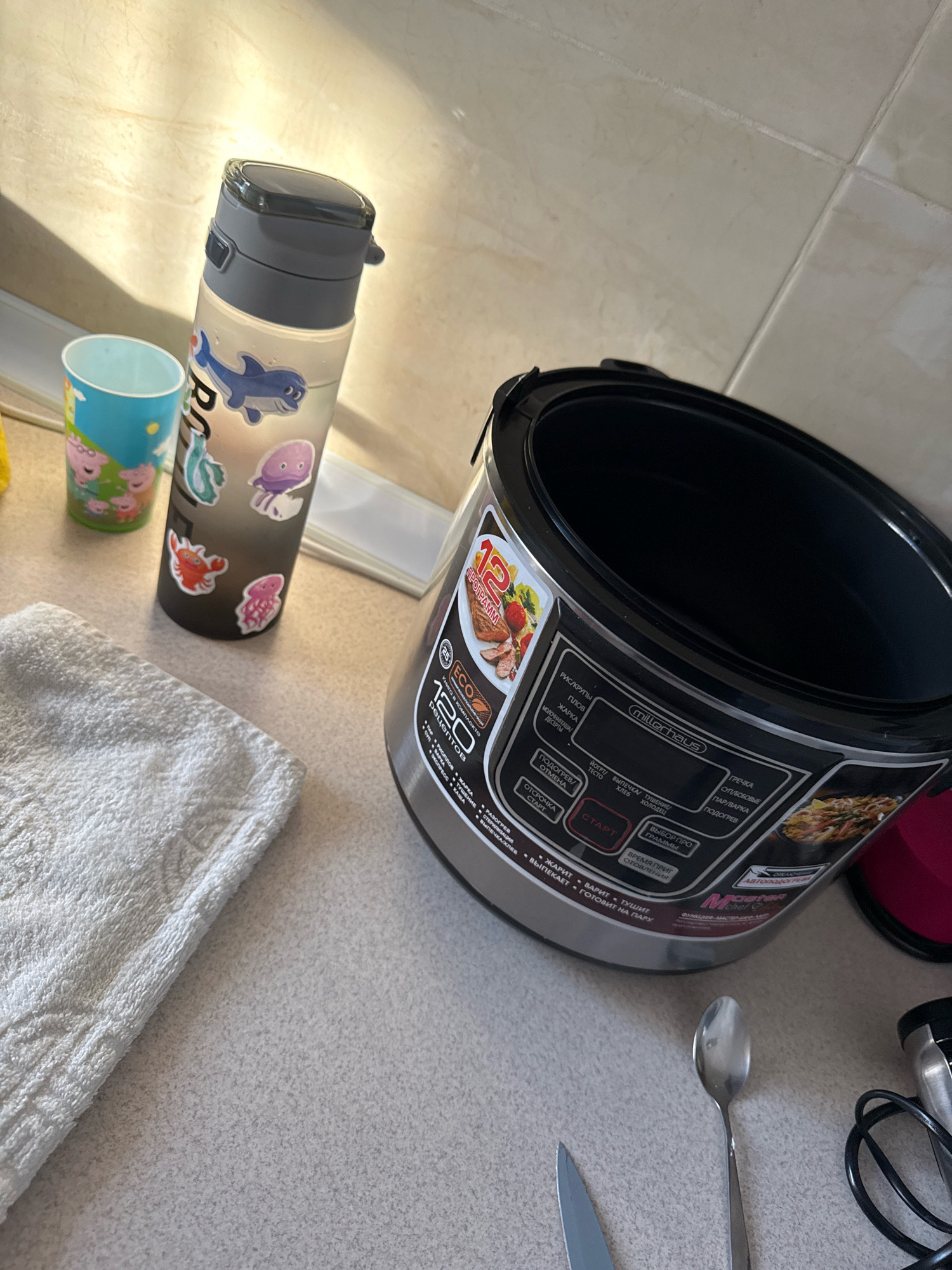 A kitchen scene features a multi-cooker, a water bottle with stickers, a cup, a folded towel, a spoon, and a knife on a countertop. The multi-cooker is silver and black with a digital display showing various cooking programs in Cyrillic script. The water bottle is clear with a grey lid and decorated with cartoon stickers of sea creatures. The cup is blue and white with images of cartoon pigs. A white terrycloth towel is folded neatly to the left of the multi-cooker. A single silver spoon and a knife are also visible on the countertop. The background consists of beige tiled walls and a light-colored countertop. The lighting suggests an indoor environment, likely a kitchen. The scene is static, with no people or active interactions depicted. There is no information in the image to indicate the location is Chișinău, Moldova.