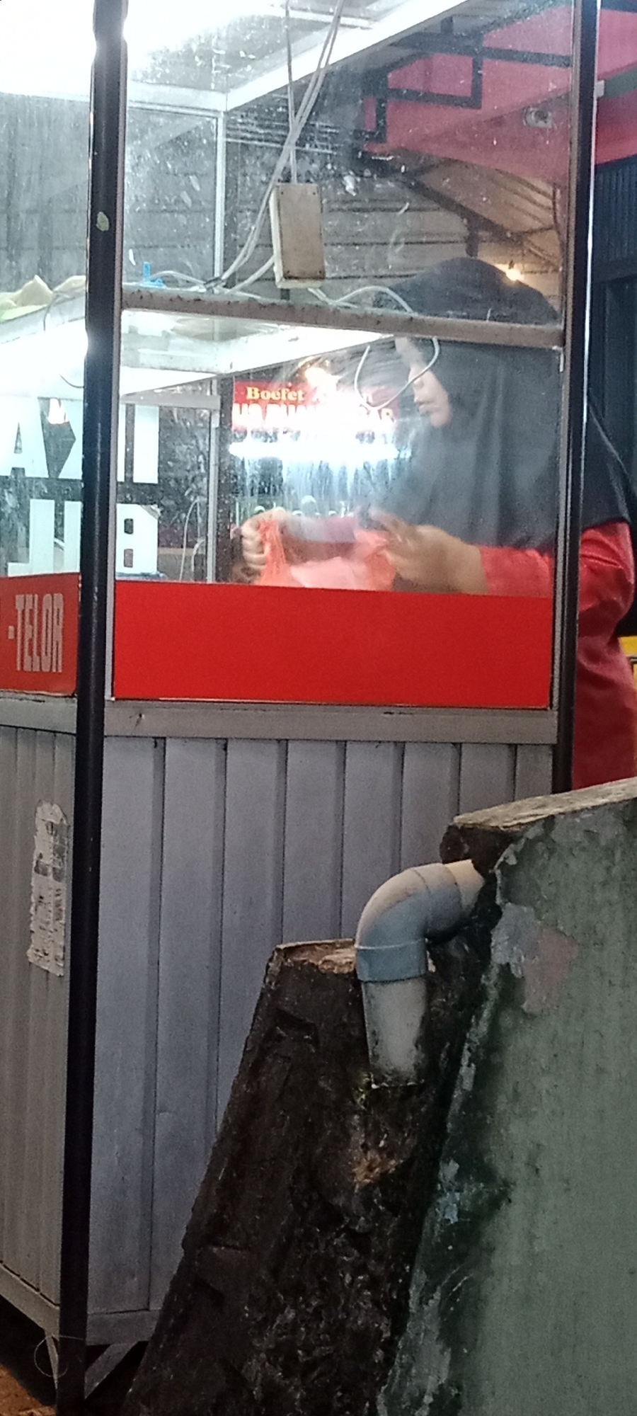 The image depicts a food stall in Tanjung Pinang, Indonesia. A woman wearing a dark hijab and a red shirt is visible behind a glass counter, interacting with an orange plastic bag. The stall has a red base and a grey corrugated metal siding. A sign with white letters reading "-TELOR" is visible on the left side. The lighting suggests it is nighttime or dusk. In the foreground, a section of a concrete structure with a drainage pipe is visible.