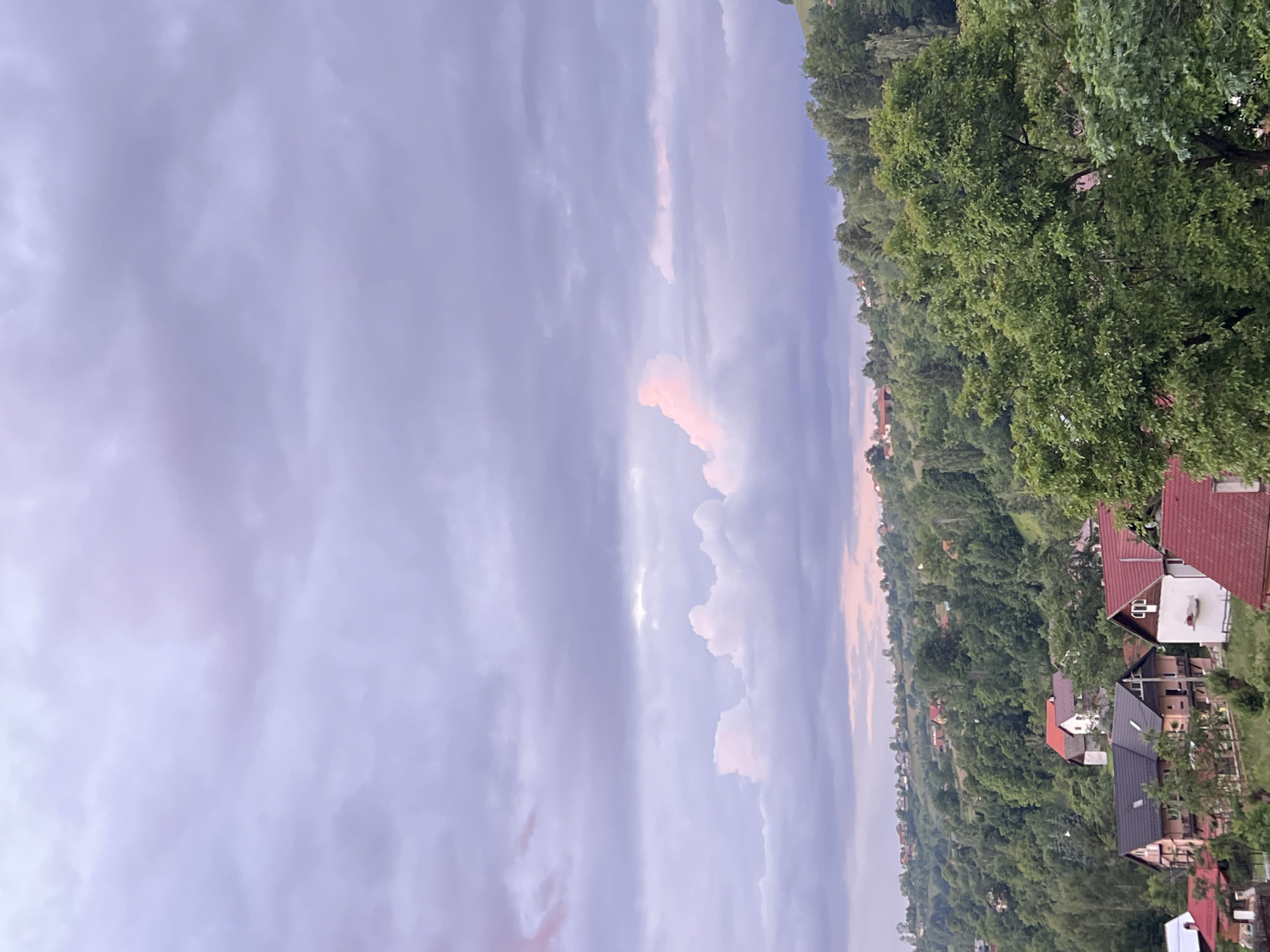The image depicts a panoramic view of a hilly, rural landscape under a cloudy, pastel-colored sky. In the foreground, several houses with red and dark-colored roofs are clustered amongst lush green trees. The houses vary in architectural style, with some featuring wooden elements and balconies. Further into the background, the landscape rises into rolling hills covered with dense forests and scattered houses, suggesting a village or small town. The sky is dominated by a mix of grey and purple clouds, with hints of pink and light blue, indicating either dawn or dusk. The overall scene conveys a sense of tranquility and natural beauty. The location is identified as being in the city of Unknown, Unknown.