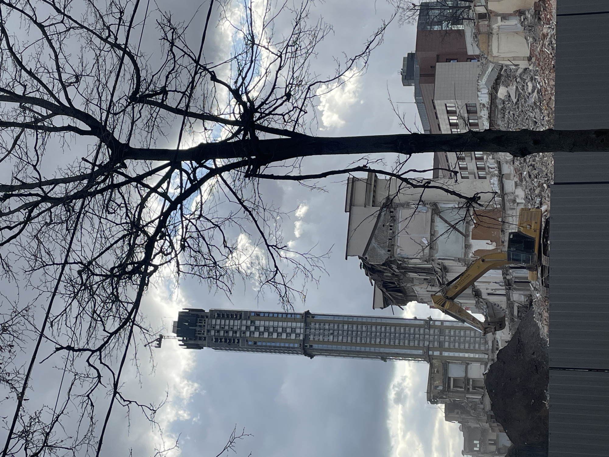 The image shows a construction or demolition site in an urban environment. A tall, slender building is under construction or renovation in the background. In the foreground, a partially demolished building is visible, with a yellow excavator actively breaking down the structure. A tree with bare branches is in the left portion of the image, silhouetted against the cloudy sky. The location is most likely Moscow, Russia.