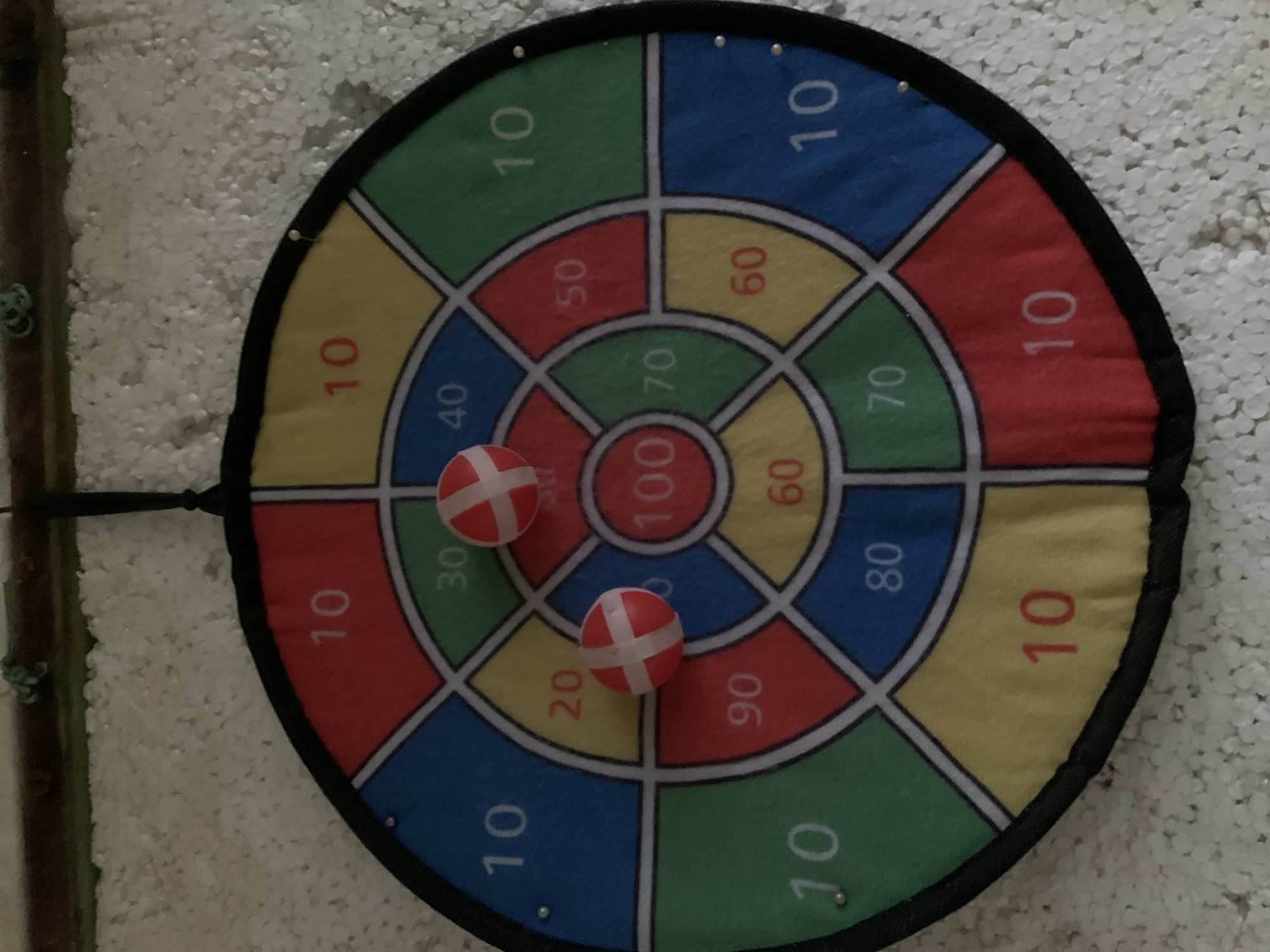 The image shows a close-up, eye-level shot of a fabric dartboard hanging on a white, textured wall, likely in an indoor setting. The dartboard is circular and divided into colored segments, each marked with a number. The numbers visible on the dartboard include 10, 20, 30, 40, 50, 60, 70, 80, 90, and 100. Two small, red and white striped balls, presumably darts, are attached to the dartboard. One ball is located in the yellow segment marked with 20, and the other is in the red segment marked with 100. A thin rope is visible at the top, suspending the dartboard. The background wall appears to be made of small, white, bead-like particles. The lighting is moderate, creating soft shadows. There are no people visible in the image. The scene appears to be in a residential or recreational space. The location in Kryvyi Rih, Ukraine, is not discernible from the visual information provided in the image.