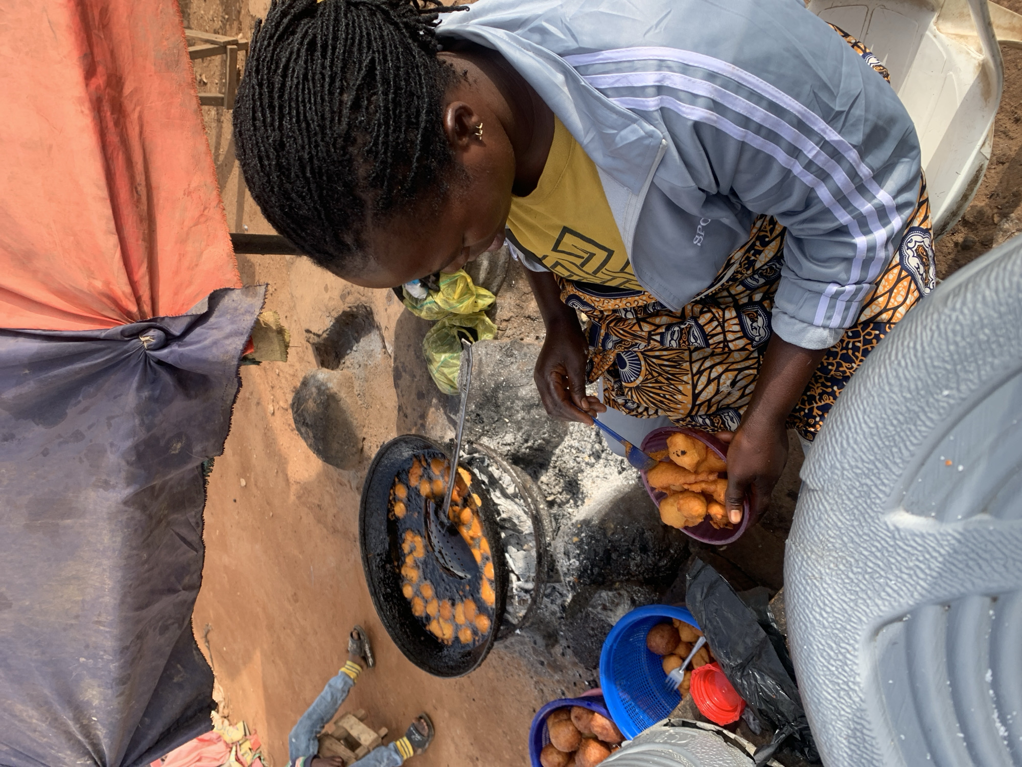 A woman with braided hair is frying dough balls in a large black pan filled with oil over a charcoal brazier. She is wearing a yellow t-shirt and a grey jacket with white stripes. She holds a purple bowl filled with fried dough balls and a serving utensil. Two blue baskets and a red plastic container are on the ground beside her. The scene takes place outdoors, possibly in Gura Top, Nigeria, with a dirt ground, some debris, and makeshift shelters visible. In the background, someone wearing blue jeans and sandals sits on a wooden structure.