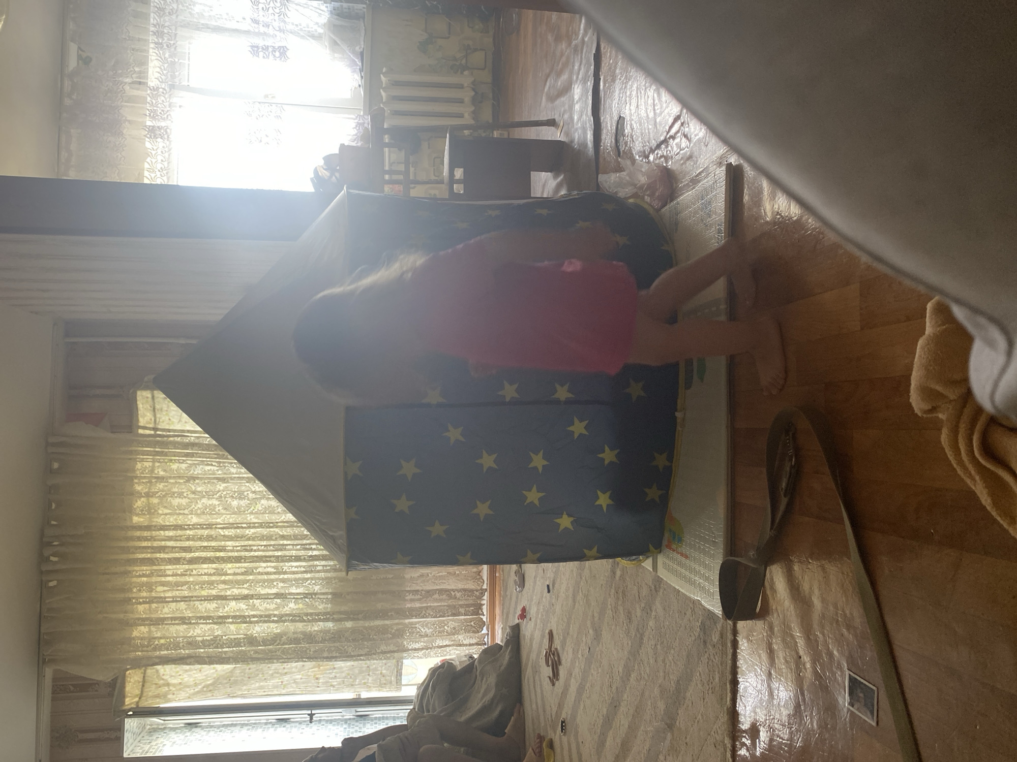 A young child wearing a pink dress is standing in a room, partially obscured by a large, blue play tent with yellow stars. The tent is positioned on a mat on a wooden floor. To the left of the tent, a grey rug is visible, and on it, a beige blanket or towel is draped over a piece of furniture. Toys, including a red object and what appears to be a pair of sandals, are scattered on the rug. A brown strap or leash is lying on the wooden floor in the foreground, with a small, dark square object nearby. Sunlight streams in from large windows covered by sheer, patterned curtains, creating a bright, somewhat hazy atmosphere in the room. The background shows a radiator and part of a chair. The scene is consistent with an interior domestic setting. Shakhtinsk, Kazakhstan, is an industrial city known for its coal mining.