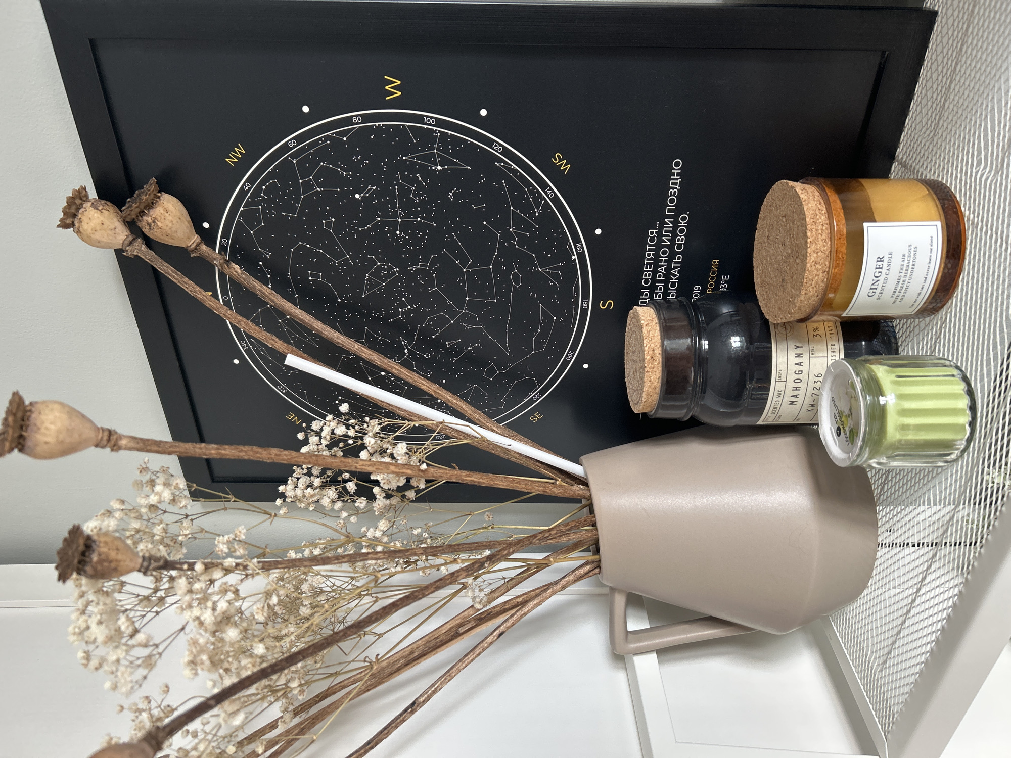 A close-up, eye-level shot captures a decorative arrangement on a white shelf. In the foreground, a small, ribbed, light green glass jar candle with a cork lid sits on a white mesh surface. To its right, a larger, amber glass jar candle with a cork lid, labeled "GINGER SCENTED CANDLE," is positioned. Behind these candles, a tall, dark brown glass jar with a cork lid and a label reading "MAHOGANY" is visible. To the left of the dark brown jar, a beige ceramic vase with a handle holds a bouquet of dried flowers and poppy seed pods. The vase is placed on the white mesh shelf. In the background, a black framed poster displaying a star chart with constellations and celestial markings is hung on a light gray wall. The poster has text in yellow and white. The environment appears to be indoors, possibly a home decor display. There are no people present in the image.