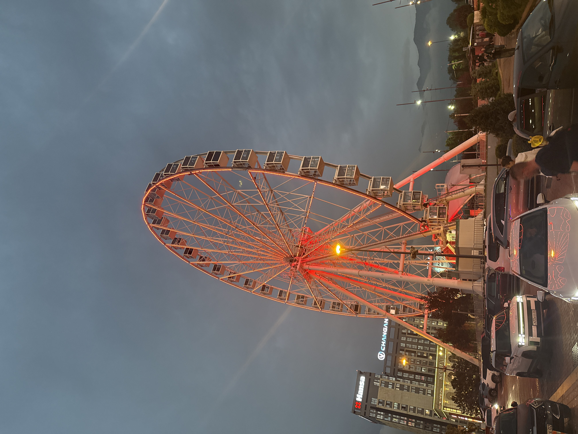 This is a nighttime photograph of a large Ferris wheel illuminated with red lights. The Ferris wheel is the dominant subject, occupying the center and right side of the frame. It is a white structure with numerous gondolas, each outlined with red lights. The wheel is situated in an area with a paved surface, suggesting a park or amusement area.
In the background, to the left of the Ferris wheel, is a tall building with the "Hansa" logo illuminated in red and white at the top. Below it, another sign reads "Miranahana Skyhall". A "CHANGAN" logo is also visible on this building. The sky is dark and overcast, with some streaks of light, possibly from airplanes or atmospheric conditions. In the far background, the silhouettes of mountains are visible, partially obscured by clouds.
In the foreground and midground, there is a parking lot filled with various cars. Several individuals are visible walking around the parking lot and near the Ferris wheel entrance. One person in the foreground is wearing a bright yellow hat.
The overall scene captures a lively evening atmosphere in what appears to be an entertainment district within the city of Almaty, Kazakhstan, given the presence of the "Hansa" building, which is a known shopping center in Almaty, and the distinct mountain backdrop characteristic of the city.