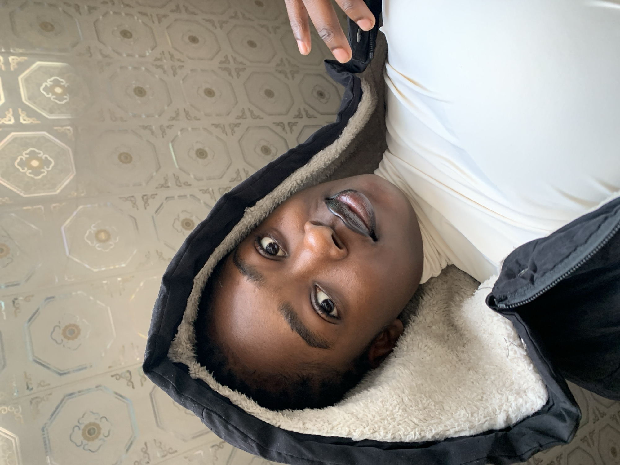This is a portrait photograph of a young Black person, likely a teenager, taken from a slightly low angle. The person is wearing a black jacket with a fuzzy, cream-colored lining, and a white garment underneath. Their head is tilted to the side, and they are looking directly at the camera with a neutral expression. The background appears to be a tiled floor with an octagonal pattern and floral motifs. The lighting suggests it is daytime. The image was taken in Jos, Nigeria.