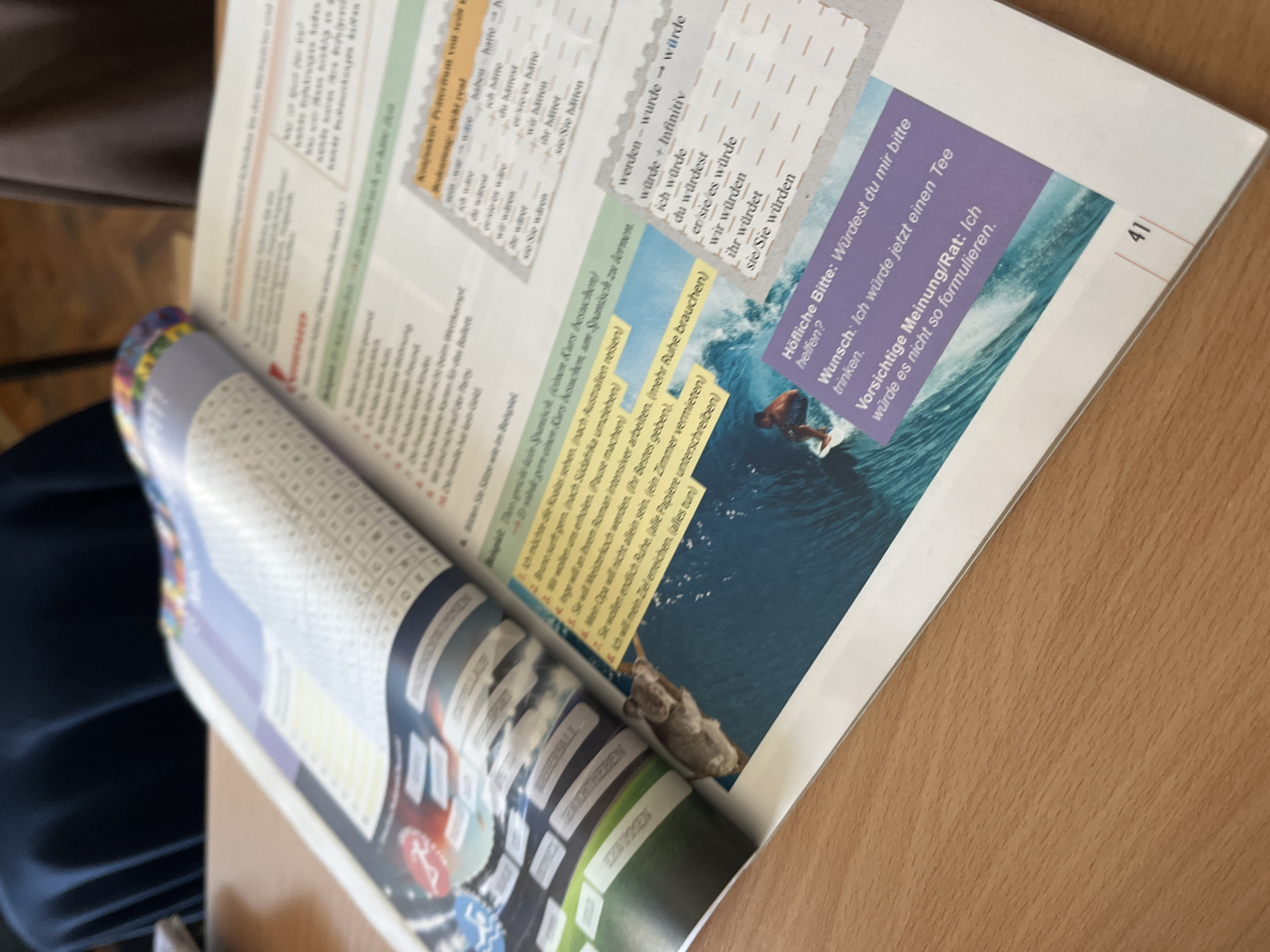 A partially curled-open book lies on a wooden surface. The pages visible contain German text and grammar exercises, with a block of text and an image of a surfer on a wave. The right-hand page is numbered "41."