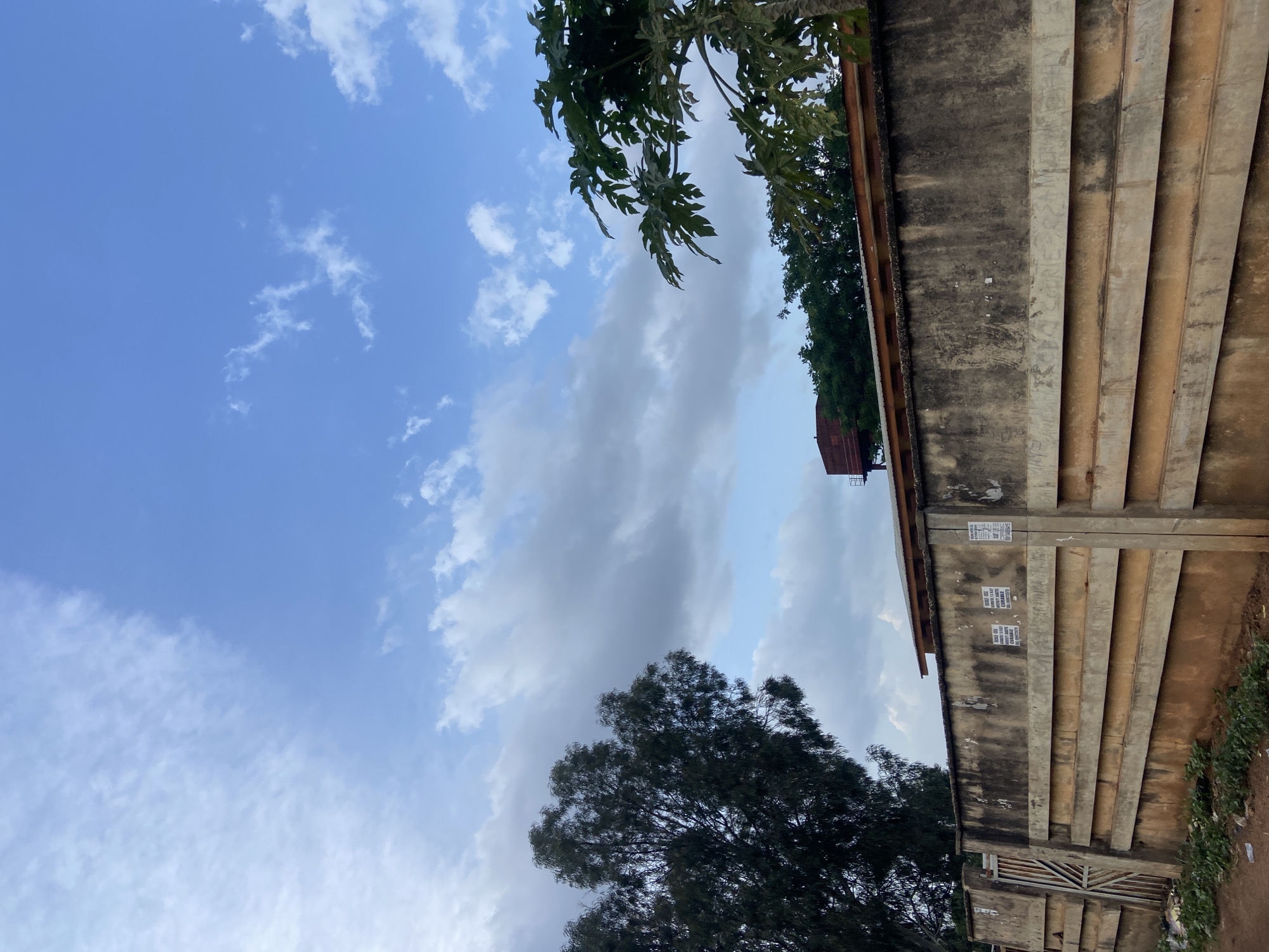 The image shows a concrete wall in Jos, Nigeria. The wall is weathered and stained, with some posters attached to it. Behind the wall, there are trees and a building with a water tower. The sky is cloudy.