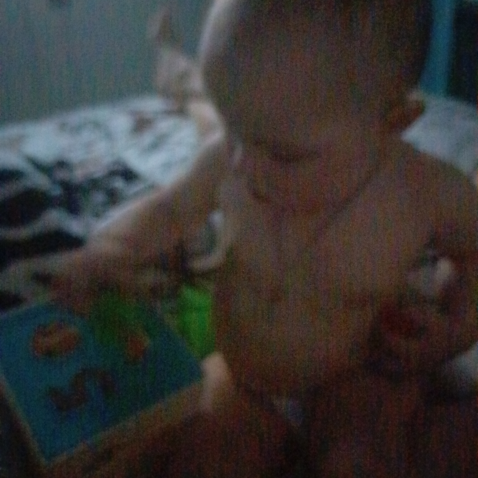 A blurry image shows a child playing with a book or learning toy in an indoor setting. The child appears to be partially undressed, with the focus on their torso and hands. A bed or sofa with patterned bedding is visible in the background. The location is unspecified.