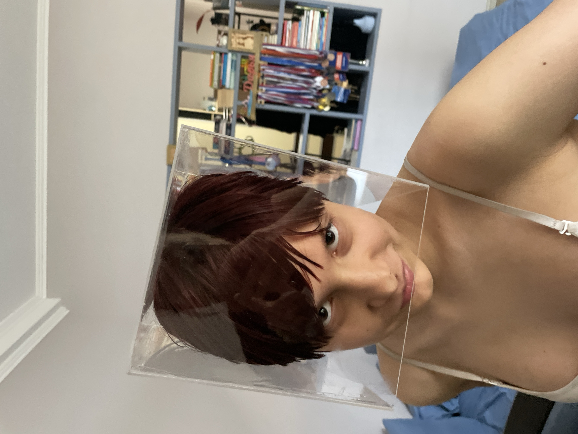 The image shows a person with reddish-brown hair, wearing a light-colored bra, looking directly at the camera. Their head is partially encased in a clear plastic cube or box. The background includes a wall with molding, a shelf unit filled with books and medals, and blue bedding. The scene appears to be indoors, possibly a bedroom. There is no indication of the location being the city of Sochi, Russia, and the description of the image is limited to the visual elements present.