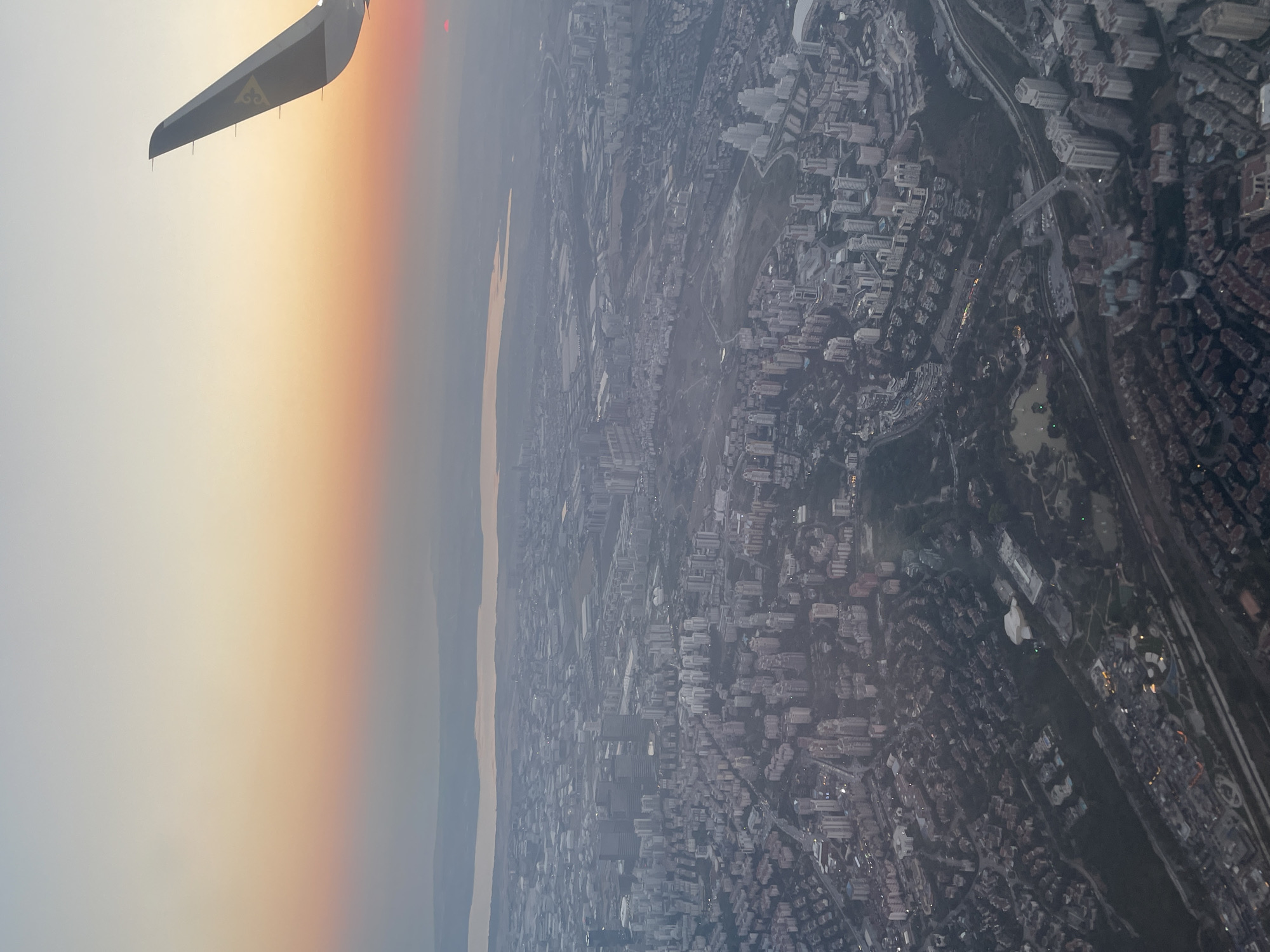 An aerial view of a dense urban landscape at sunset is captured from an airplane window. The sky transitions from a pale, hazy yellow at the top to soft oranges and pinks closer to the horizon. The sun, a small red disc, is visible on the right edge of the frame, partially obscured by haze.
In the foreground, an airplane wing, dark gray with a yellow logo, extends into the upper right corner of the image. Below the wing, a vast city unfolds, characterized by numerous buildings of varying heights and architectural styles. Tall, modern skyscrapers, many with white facades, are clustered together, interspersed with lower-rise residential buildings and a sprawling network of streets and roads.
Further into the landscape, a body of water, likely a river or strait, is visible in the mid-ground, reflecting the muted colors of the sky. The shoreline is lined with more urban development. The overall scene suggests a densely populated metropolitan area experiencing the transition from day to night, with the warm glow of sunset illuminating the urban sprawl. The city appears to be Beykoz, Turkey, given the topography and urban planning visible. There are no people or specific actions discernible in the image.