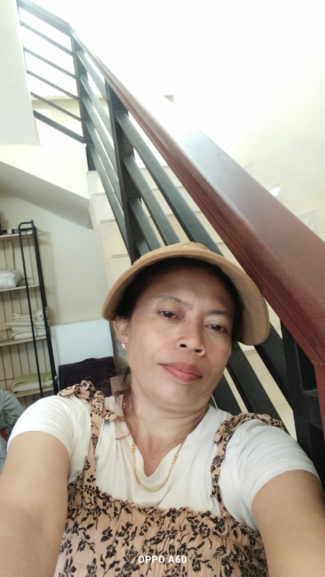 An overhead, close-up, selfie-style shot shows an adult Asian woman looking directly at the camera. She is wearing a tan-colored hat, a white t-shirt, and a patterned sundress. She has brown eyes, a light tan complexion, and is wearing light makeup. Her hair is dark brown and pulled back. A thin gold necklace is visible around her neck. The woman is seated on a staircase, with her body angled away from the camera to her right. The background consists of a dark brown wooden handrail and a black metal railing, which are part of an indoor staircase. The walls are painted white. To the left of the woman, a metal shelving unit with items on it is visible. The setting appears to be a residential interior, possibly in Denpasar, Indonesia, given the "OPPO A60" watermark on the image, which is a mobile phone model often sold in Southeast Asia. The lighting suggests an indoor setting with overhead lighting or natural light from a nearby window.