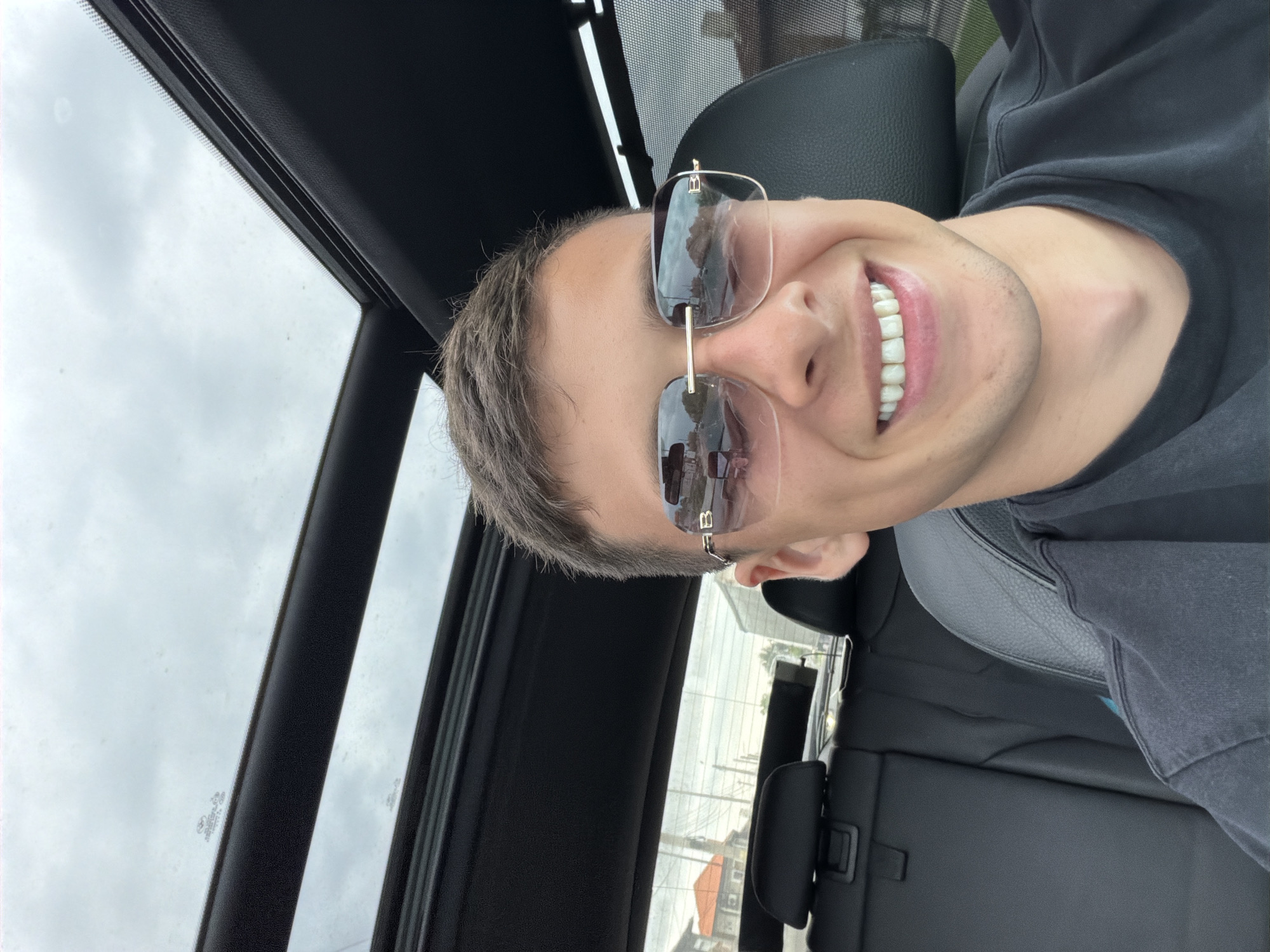 A portrait-oriented image, rotated 90 degrees counter-clockwise, depicts a young man taking a selfie from inside a car. The man, with short, dark hair, is the primary subject, positioned in the foreground and center. He is wearing rectangular, gold-framed sunglasses, possibly with a "B" or "FB" logo visible on the temple arm. He has a wide, cheerful smile, showing his teeth.
He appears to be seated in the driver's or front passenger's seat, with a black leather car seat visible behind him and to his side. The car's interior trim, predominantly dark grey or black, is seen around him. Above him, a sunroof reveals an overcast, cloudy sky. Through the car's windows, a grey, cloudy sky is visible, along with reflections that show parts of buildings, utility poles, and wires, suggesting an outdoor, possibly urban or suburban, setting. A notable detail is a Mercedes-Benz logo etched into one of the side windows. The overall impression is one of happiness or contentment, captured in a casual moment.