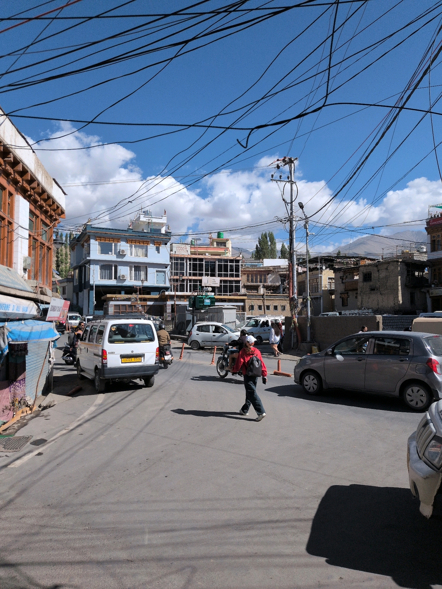 In the city of Leh, India, a narrow asphalt street is crowded with vehicles and pedestrians. A white van, possibly a taxi due to its license plate, is parked on the right side of the road. A young person in a red jacket and dark pants is walking across the street from left to right, carrying a black backpack. A gray hatchback car is parked on the right side of the street, and a motorcycle with a rider is approaching from the background. Several other vehicles, including a white van and a scooter, are visible in the background. The street is lined with multi-story buildings, some of which are shops or hotels. A prominent feature of the scene is the dense network of overhead electrical wires, which crisscross the sky. The sky is a vibrant blue with scattered white clouds. The overall impression is one of a bustling urban environment in a mountainous region.