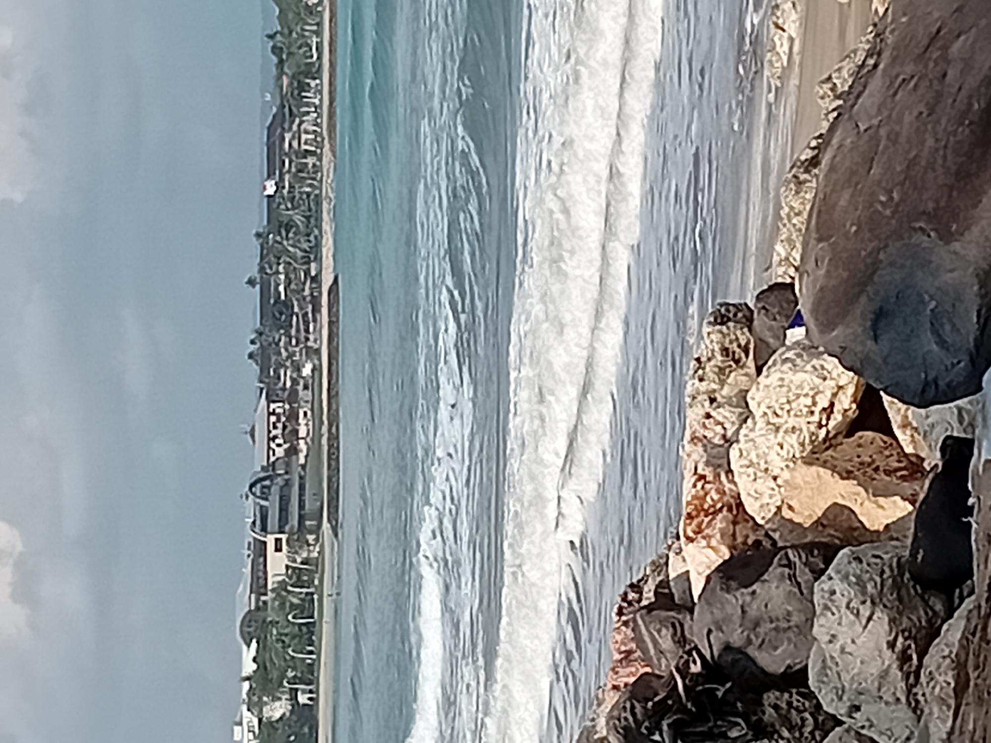 The image depicts a coastal scene in Kuta, Indonesia, with a focus on the ocean and a rocky shoreline. In the foreground, a pile of large, irregularly shaped rocks, some appearing wet and dark, others lighter and sandy in color, are visible. Some of these rocks are partially covered by seaweed or other organic debris.
Beyond the rocks, the ocean stretches out with a series of gentle waves breaking and forming white foam. The water is a cool blue-green, with varying shades indicating depth and light. In the middle ground, a sandy beach is visible, with a faint line of the surf washing ashore.
The background reveals the coastline of Kuta, characterized by a strip of buildings and palm trees. These structures appear to be hotels or resorts, with modern architectural designs featuring curved roofs and multiple stories. The buildings are situated along the beach, interspersed with lush green foliage. The sky above is overcast with a mix of grey and white clouds, suggesting a partly cloudy day. A faint American flag is visible on one of the buildings in the distance, and a few indistinct figures, likely people, can be seen on the beach in the far distance. The overall impression is of a tropical beach resort setting.