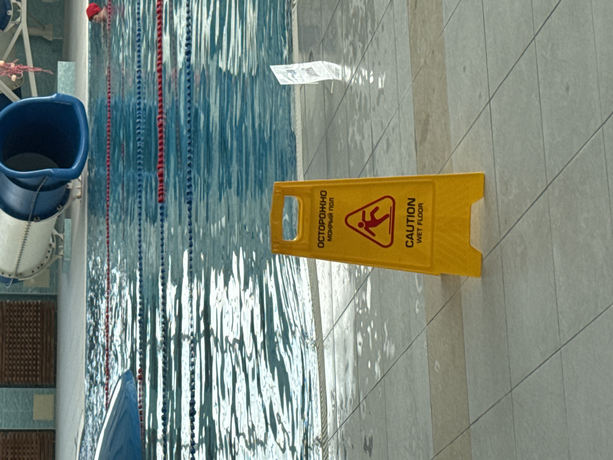 The image depicts an indoor swimming pool area. In the foreground, a yellow "caution wet floor" sign is prominently displayed on the tiled floor. The sign is in English and Russian, with the Russian text reading "ОСТОРОЖНО МОКРЫЙ ПОЛ". The floor around the sign is wet and tiled. In the mid-ground, the swimming pool itself is visible, with lane ropes dividing the water. A blue slide structure and part of a white water slide are visible in the background. To the right, a person wearing a red swim cap is partially submerged in the pool. The overall environment appears to be a public leisure facility. Based on the Russian text on the sign, the location is likely within the city of Perm, Russia.