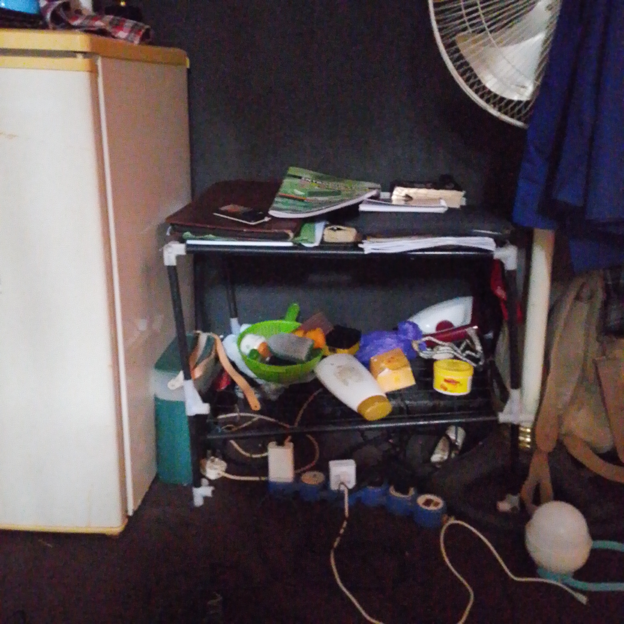 The image depicts a cluttered indoor scene, likely a living space or storage area, in Jalingo, Nigeria. A white refrigerator stands to the left, its surface partially obscured by what appears to be a checkered cloth. Next to it, a black metal shelf unit is overloaded with various items.
The top shelf holds a stack of papers, some appearing to be magazines or notebooks, with one green and white publication prominently displayed. A dark-colored binder or book is also visible, alongside a small, light-colored object.
The second shelf is filled with an assortment of objects. A bright green bowl, possibly containing cleaning supplies or small items, sits towards the left. Adjacent to it are various bottles and containers, including a white bottle with a yellow cap, a purple item, and a small yellow container. Some wires and cables are also visible, trailing from objects on this shelf and onto the floor.
The bottom of the shelf unit and the floor are also cluttered with more items and cables. A teal-colored container is positioned to the left of the shelf. Towards the right side of the image, a white fan with a circular grill is partially visible against a dark wall. A portion of dark blue fabric, possibly clothing or a curtain, hangs to the right of the fan. A beige bag or backpack rests on the floor to the far right. A white spherical object, possibly a light fixture, sits on the floor near some blue tape.
The lighting suggests it is daytime, but the indoor setting and the presence of a fan indicate potentially warm weather. The overall impression is one of a lived-in and somewhat disorganized space. No people are visible in the image. No discernible text is legible.