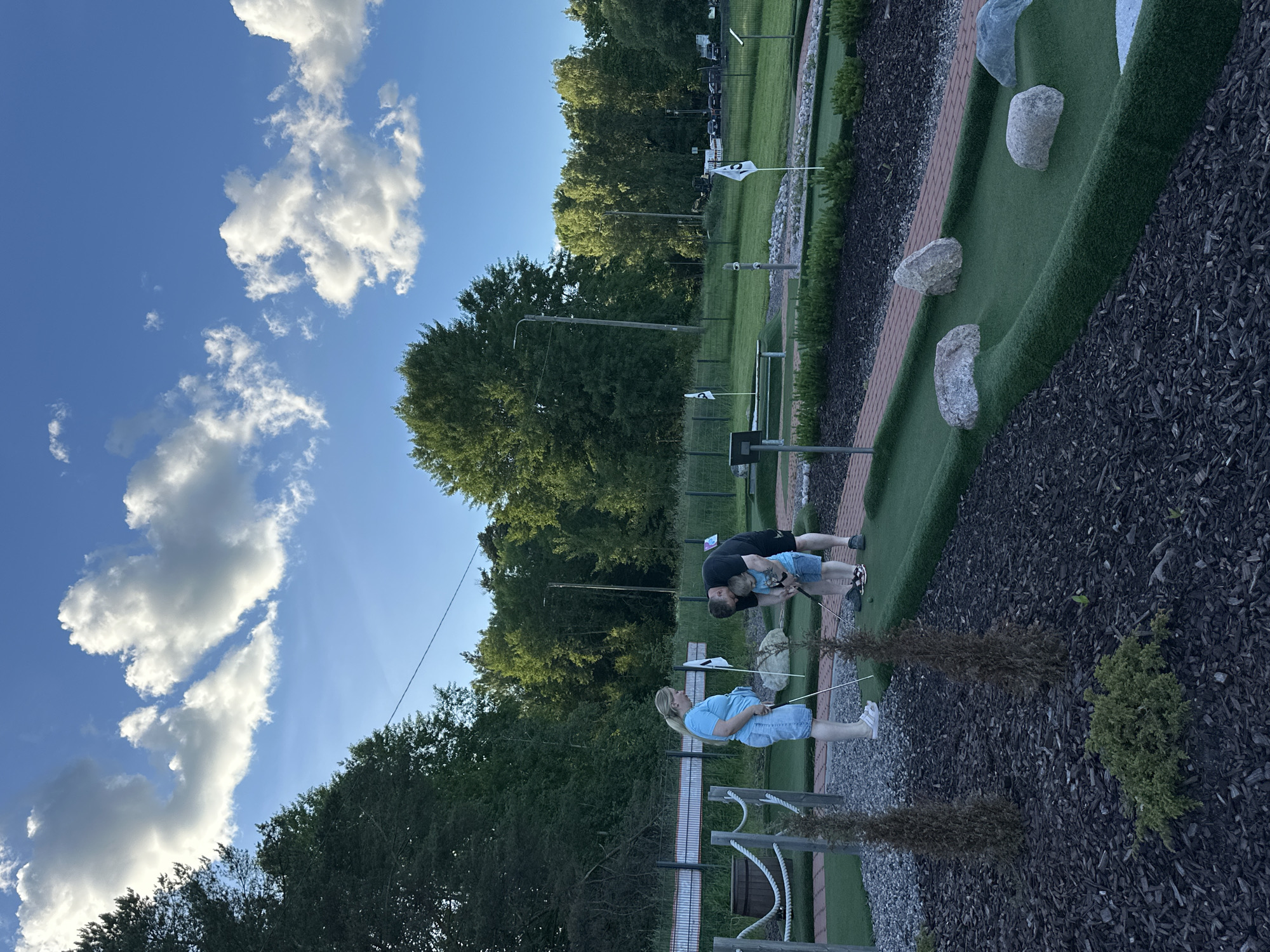 In an outdoor mini-golf course, a family is playing. A man is kneeling and assisting a young boy in swinging a putter, while a woman stands nearby holding a putter. The course features artificial turf greens, brick borders, and strategically placed rocks as obstacles. The background consists of lush green trees and a clear blue sky with scattered clouds. The scene appears to be set in a park or recreational area.