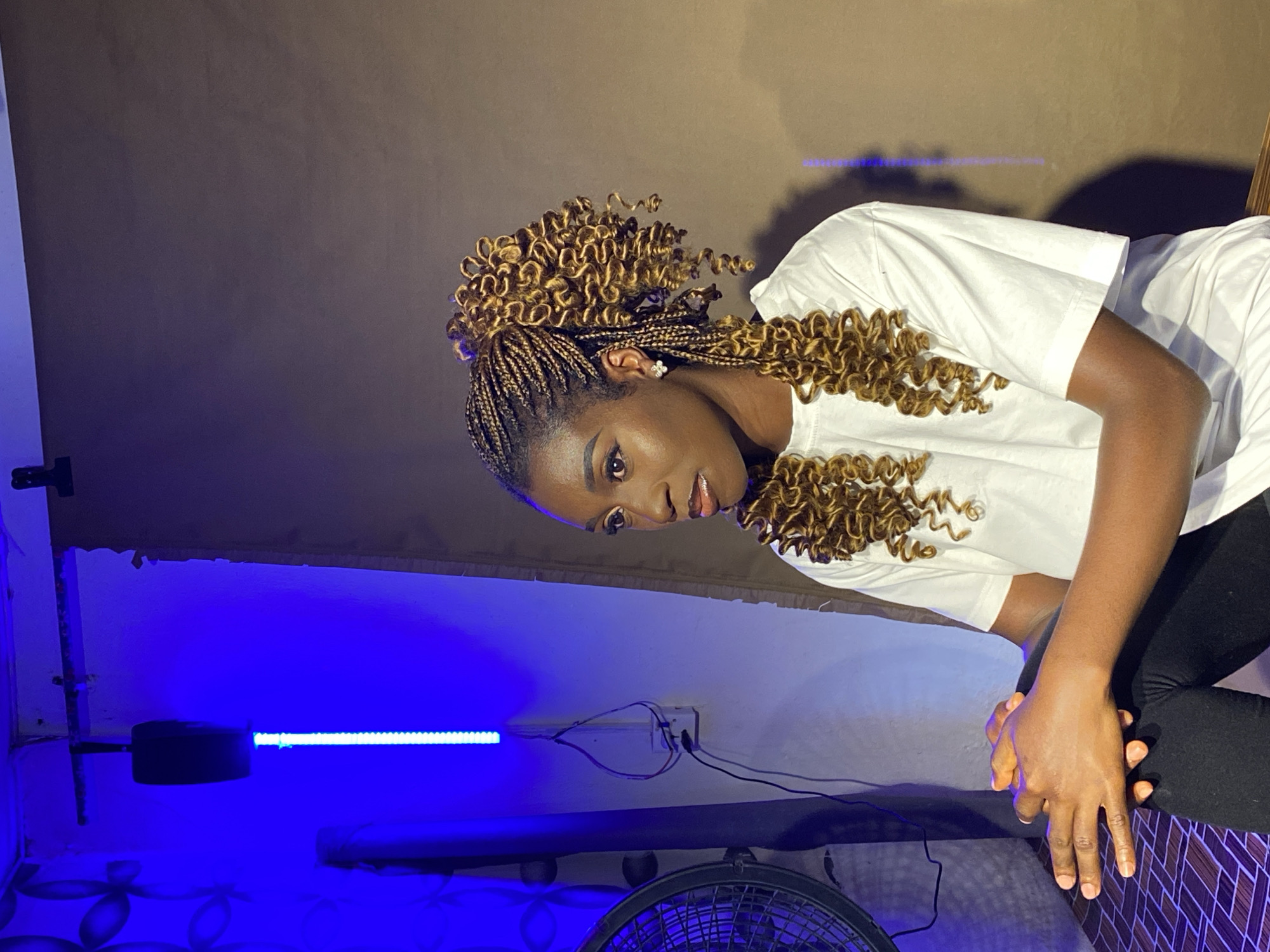 A woman with braided brown hair, styled with curly ends, wears a white t-shirt and dark leggings. She is leaning forward, hands clasped together. The background is two-toned, with a brown upper section and a blue lower section, illuminated by a horizontal blue light. A black fan is visible in the foreground. The location is Vom, Nigeria.