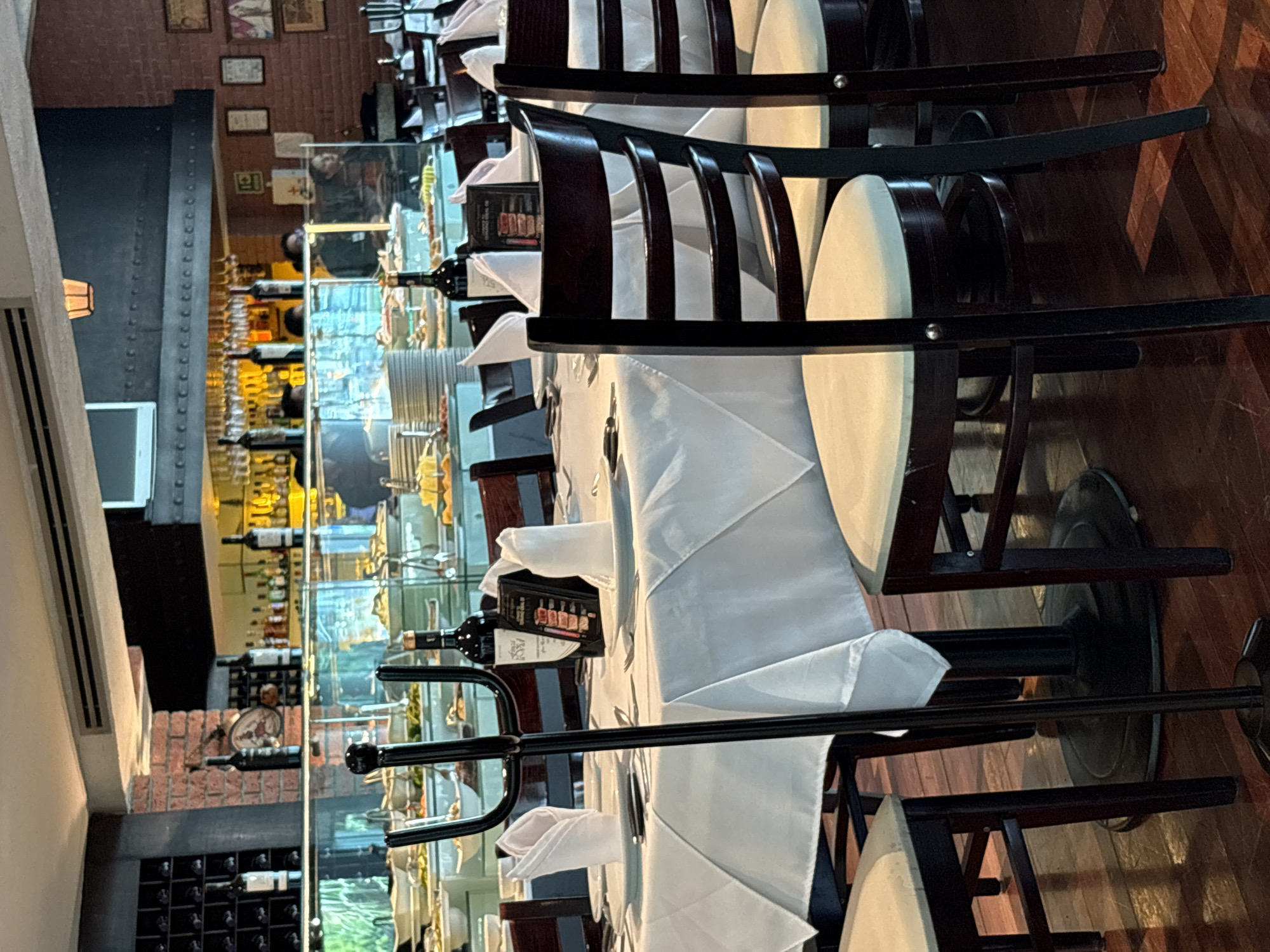 The image depicts a high-end restaurant interior, likely in Huixquilucan de Degollado, Mexico. The scene is dominated by a long, formally set table covered with a white tablecloth and napkins, surrounded by dark wood chairs with white cushioned seats. Wine bottles and menus are placed on the table, suggesting a fine dining experience. In the background, a display case houses numerous wine bottles and glassware. A brick wall and a black metallic structural element are also visible. The overall impression is of a sophisticated and upscale establishment.