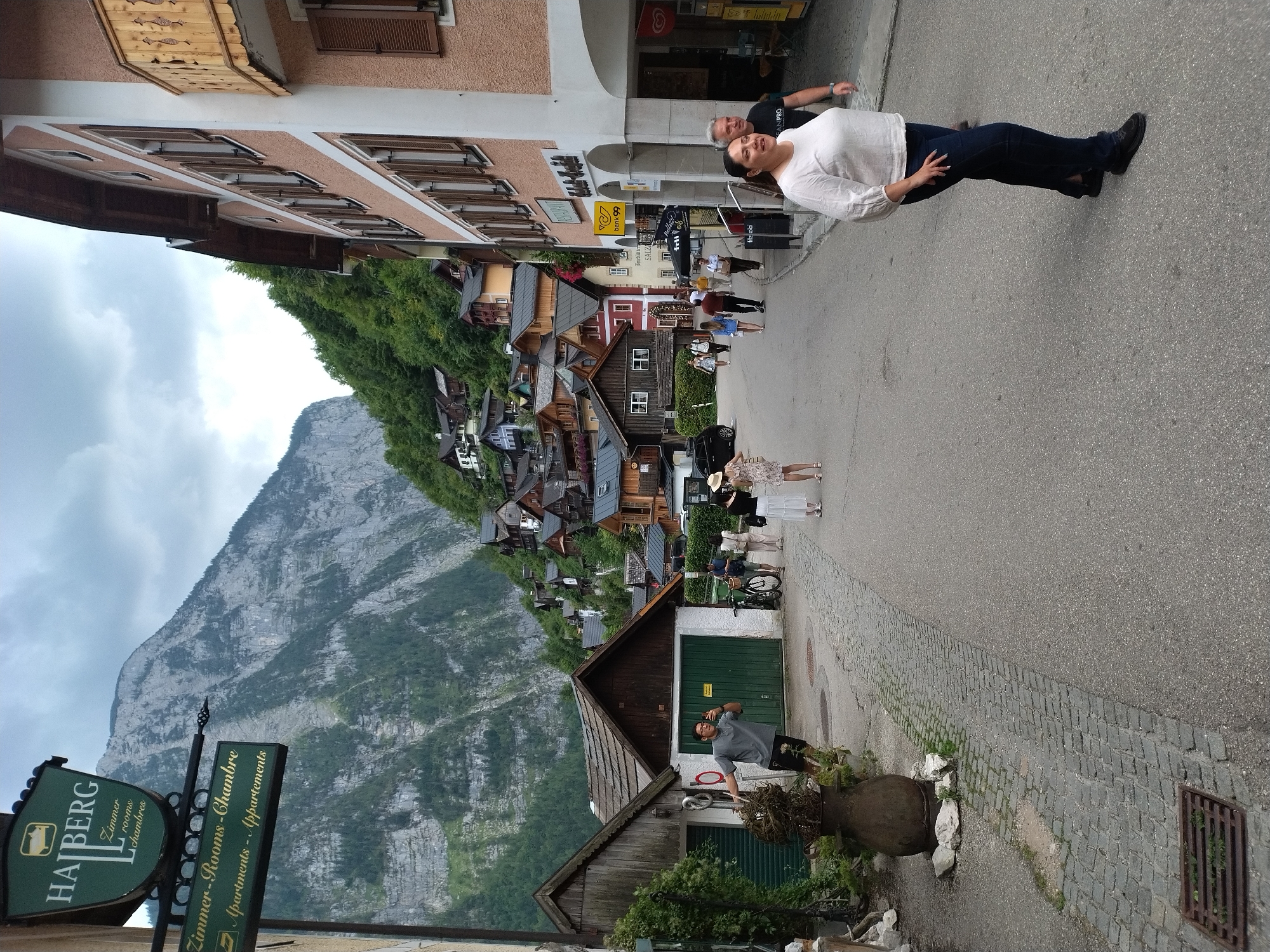 This image captures a street scene in Hallstatt, Austria, with traditional Alpine architecture dominating the background. The iconic Hallstatt mountain rises steeply behind the village, its rugged slopes covered in lush green trees. A cluster of wooden houses with dark roofs cascades down the hillside, a quintessential image of this UNESCO World Heritage site.
In the foreground, a paved street runs through the village. On the left, a sign for "HAUBERG" is visible, indicating lodging services with "Zimmer - rooms - chambres" and "Apartments - appartements" advertised below. A man in a grey t-shirt and black shorts is posing for a selfie in front of a rustic building with a green door.
Further down the street, several people are walking. A couple in white is seen on the right side of the street, and other individuals are scattered throughout the scene, some walking in groups, others alone. The architecture features pastel-colored buildings with wooden shutters and balconies, typical of the region. The street is lined with cobblestones in some areas and asphalt in others. The overall atmosphere suggests a tourist-friendly environment on a cloudy day.