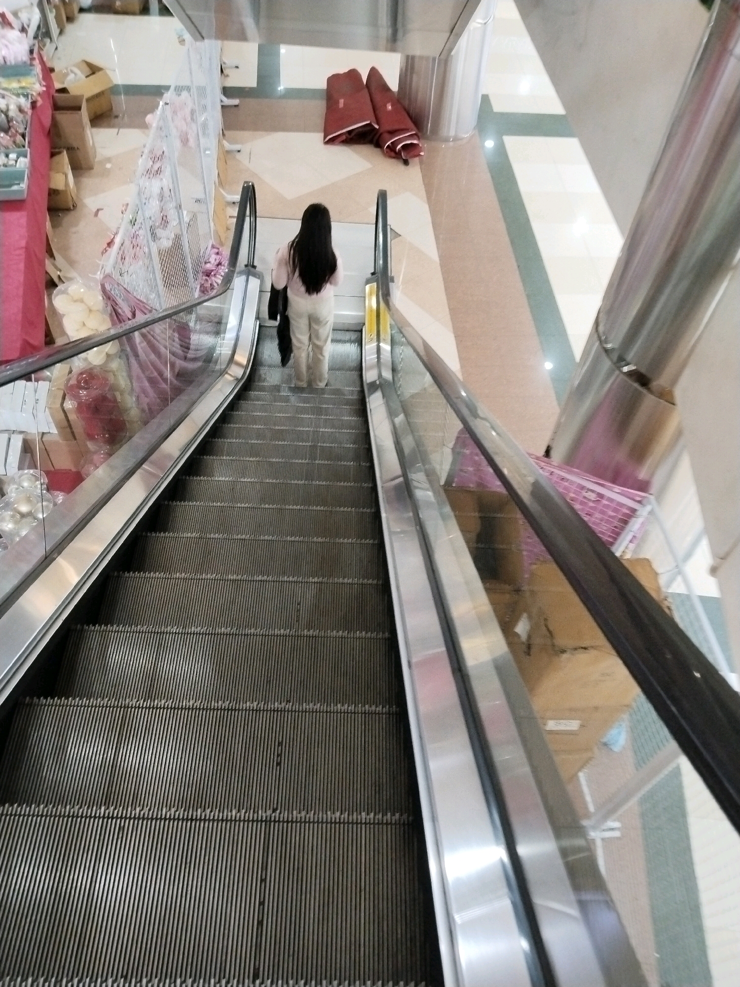 A person with long dark hair, wearing a light pink long-sleeved shirt and beige pants, stands on a downward-moving escalator in a shopping mall. They are holding a dark bag in their right hand. The escalator has metal steps and glass and metal railings. On the left side of the escalator, there are displays of various items, including boxes, bags, and decorative spheres. On the right side, a large metallic cylindrical structure is visible, and further back, there are folded red fabrics and a patterned tiled floor. The environment suggests a commercial setting, likely a mall in Surabaya, Indonesia, given the provided context, with an upward view from the escalator.