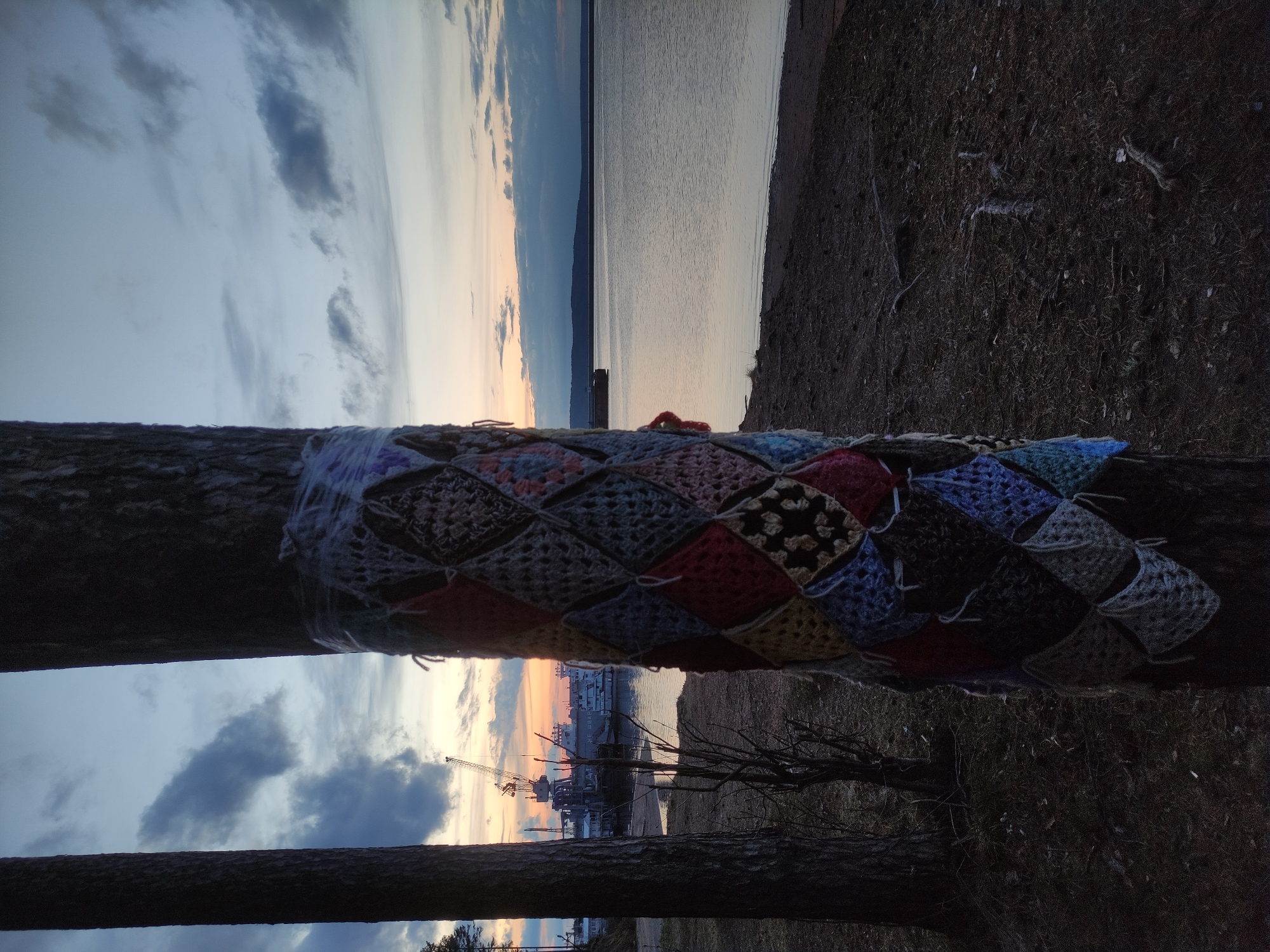 The image depicts a serene landscape at what appears to be dusk or dawn, showcasing a contrast between natural elements and industrial activity. In the foreground, two prominent tree trunks frame the scene vertically. The left tree trunk is adorned with a colorful "yarn bomb" or "tree cozy" made of crocheted granny squares in various hues of red, blue, green, yellow, brown, and grey. The ground to the right of the trees consists of dark, sparse earth or vegetation.
Beyond the trees, a wide body of calm water, likely a river or reservoir, stretches horizontally across the midground. On the far bank, industrial infrastructure is visible, including several large cranes and buildings, suggesting a port or shipyard. A large vessel, possibly a ship or barge, is docked at this facility, with some Cyrillic text, potentially part of its name, visible on its side, starting with "НЕ..." (NE...).
The background features a vast sky transitioning from warm hues of orange and yellow near the horizon, indicative of sunrise or sunset, to cooler blues and greys higher up. Scattered clouds, predominantly dark grey, are spread across the sky. The overall lighting is soft and atmospheric, casting a tranquil mood over the scene despite the industrial presence. The location context is Chaikovsky, Russia. No specific human activity is depicted, but the yarn bomb implies human presence and creative expression.