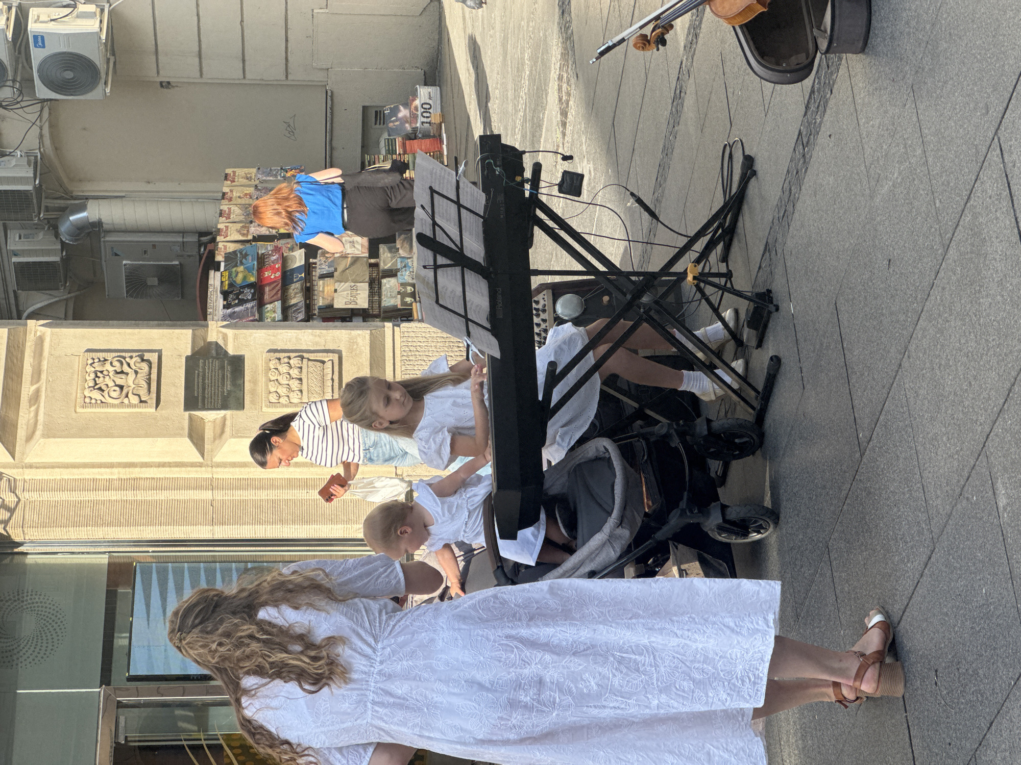 This media file captures a lively street scene in Belgrade, Serbia, on a sunny day. The primary focus is on a group of people, seemingly a family, engaged in a musical performance. A woman with long, blonde, wavy hair, wearing a white embroidered dress, stands in the foreground, partially obscuring a baby in a stroller. Two young girls, one with blonde hair and the other with darker hair, are positioned near a keyboard. The blonde girl is looking at a music sheet, while the darker-haired girl is focused on the keyboard. A baby is also visible in a stroller, appearing to be part of the group.
Behind the performers, a stall displays a variety of books and possibly CDs. The backdrop is a building with architectural details and air conditioning units. A violin and its case are visible on the ground near a keyboard stand, suggesting a musical ensemble. The pavement is paved with grey stone tiles. The sunlight indicates it's daytime, likely mid-morning or afternoon, with bright shadows cast on the ground. The overall mood appears to be relaxed and cheerful, with the family enjoying their performance on the street.
Visible text includes "Roland" on the keyboard stand and "Beatles" on some of the books or CDs. The presence of a stroller and the general atmosphere suggest a family outing or a street performance.