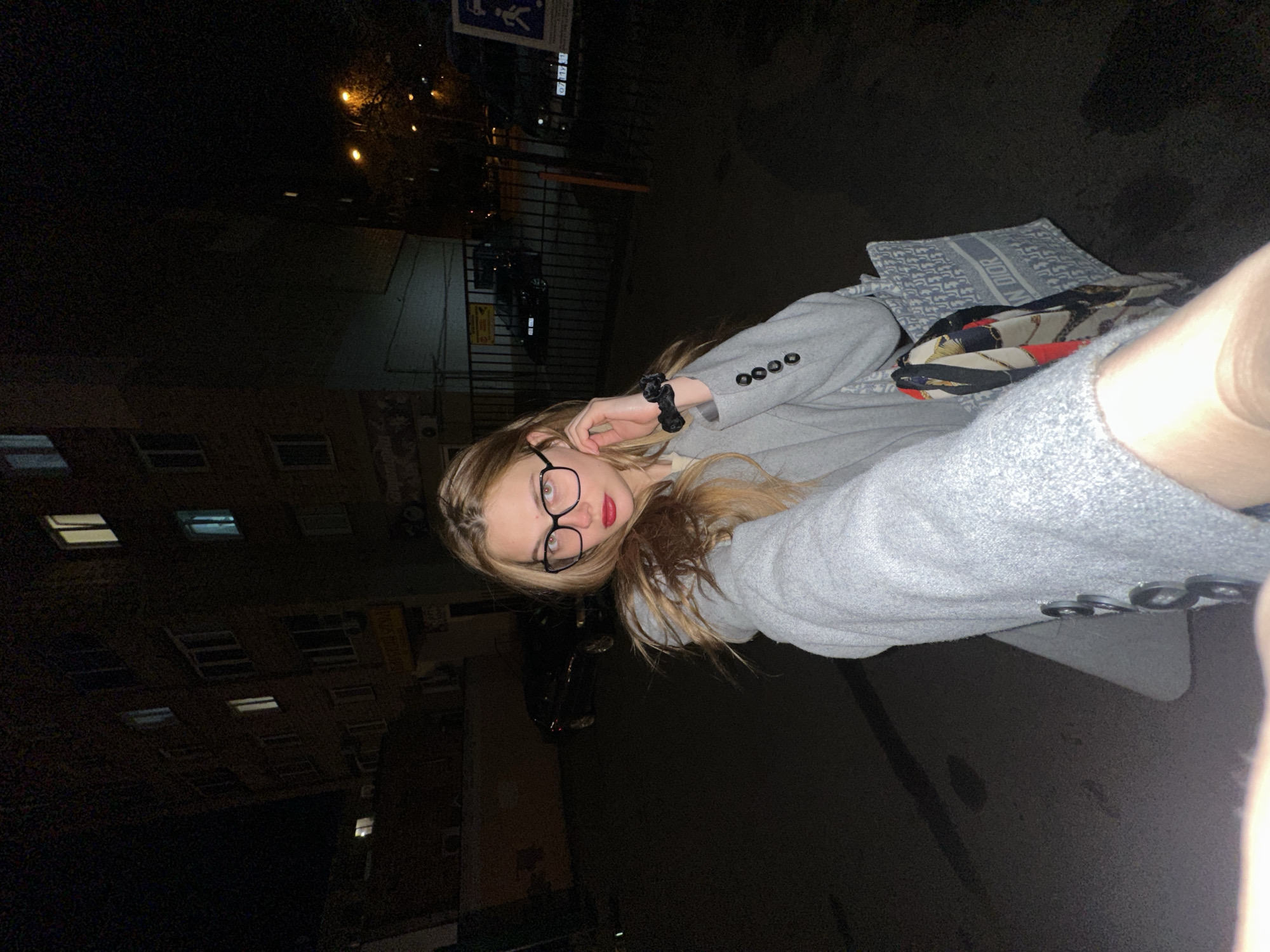 A young woman with glasses and red lipstick is shown at night. She is wearing a gray coat and sitting with a designer bag in her lap. The location is likely Krasnoyarsk, Russia, based on the buildings in the background which are typical of urban architecture in that city. The scene is dimly lit, with streetlights and building windows providing the primary sources of illumination. A car is parked nearby.