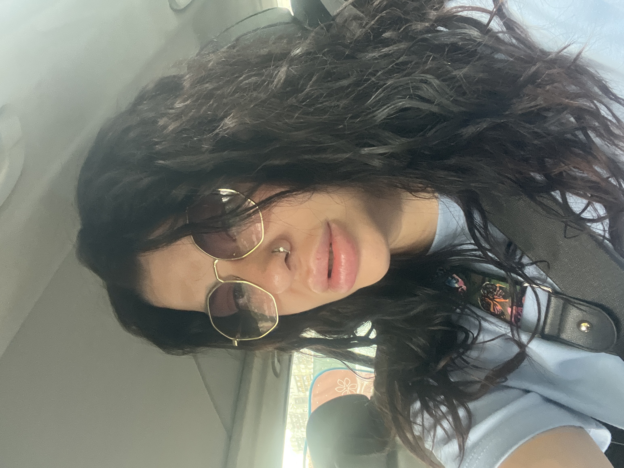 A close-up portrait shows a young person with long, voluminous dark curly hair, positioned inside a vehicle, possibly in Plovdiv, Bulgaria. The individual is wearing stylish hexagonal-framed sunglasses with gold-toned metal, obscuring their eyes. A small, circular silver septum piercing is visible on their nose, and their lips are slightly parted in a neutral or subtle expression. They are dressed in a light blue t-shirt. A black strap, resembling a car seatbelt or a bag strap, crosses their shoulder and chest. Partially visible underneath the strap and strands of hair is a colorful, patterned patch or design on the t-shirt, featuring abstract shapes and hints of floral motifs.
The background consists of the car's interior, with a light-colored ceiling visible towards the upper left. Natural light streams in from outside a window, suggesting it is daytime, with blurred shapes and bright light discernible in the background. The subject is looking directly towards the camera, indicating a selfie or a casual photo opportunity. No visible text is present in the image.