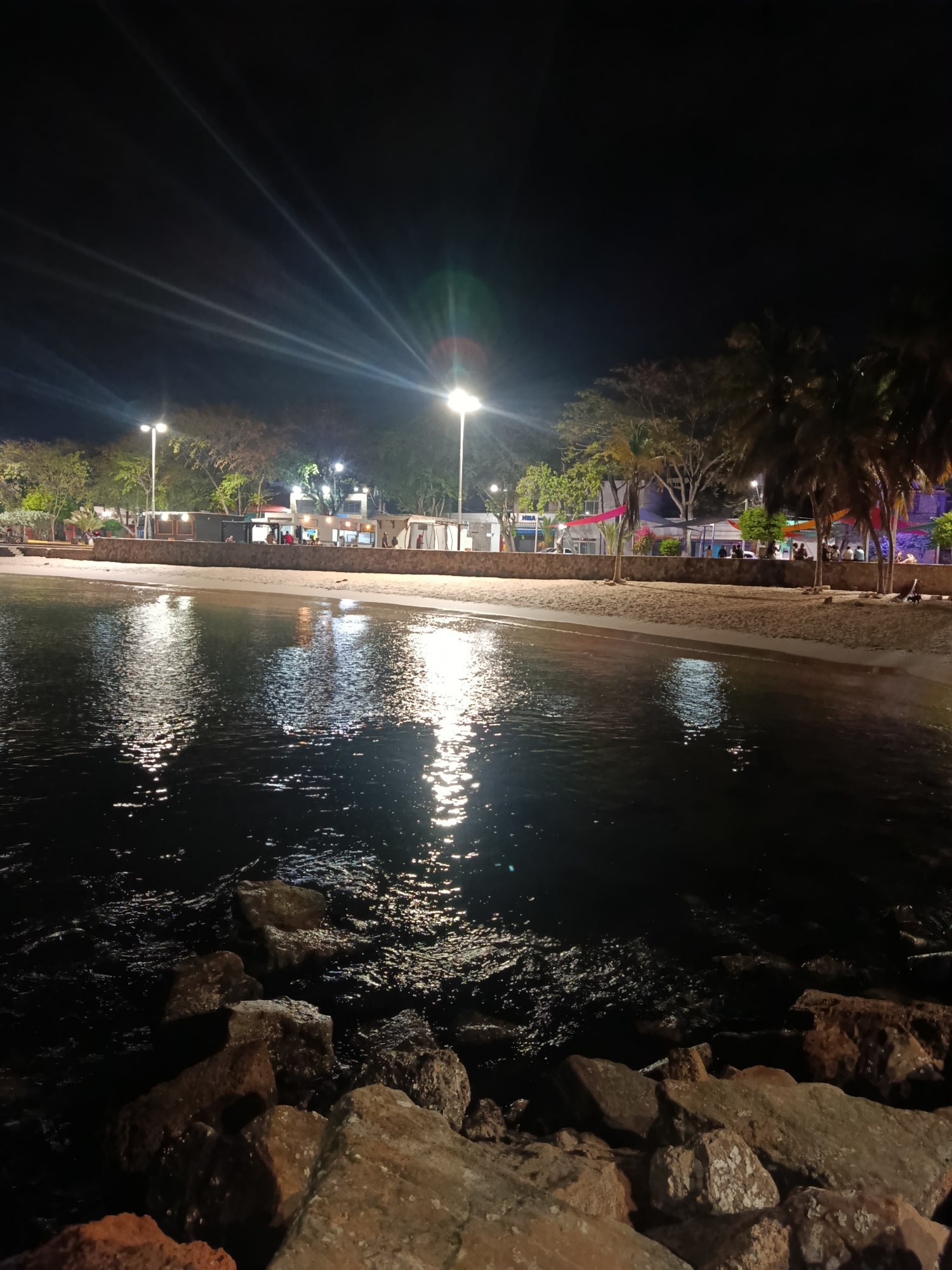 This nighttime image depicts a waterfront scene in Juan Griego, Venezuela. In the foreground, large rocks form a barrier, with water lapping around them. The midground features a sandy beach leading to a waterfront promenade, lined with several small buildings and structures lit by streetlights. People are visible along the promenade, though details are unclear due to the distance and lighting. Palm trees are interspersed along the beach and promenade, adding to the coastal atmosphere. The background is dark, punctuated by the glow of streetlights and illuminated buildings. The water reflects the artificial light sources, creating bright streaks and patterns across its surface.