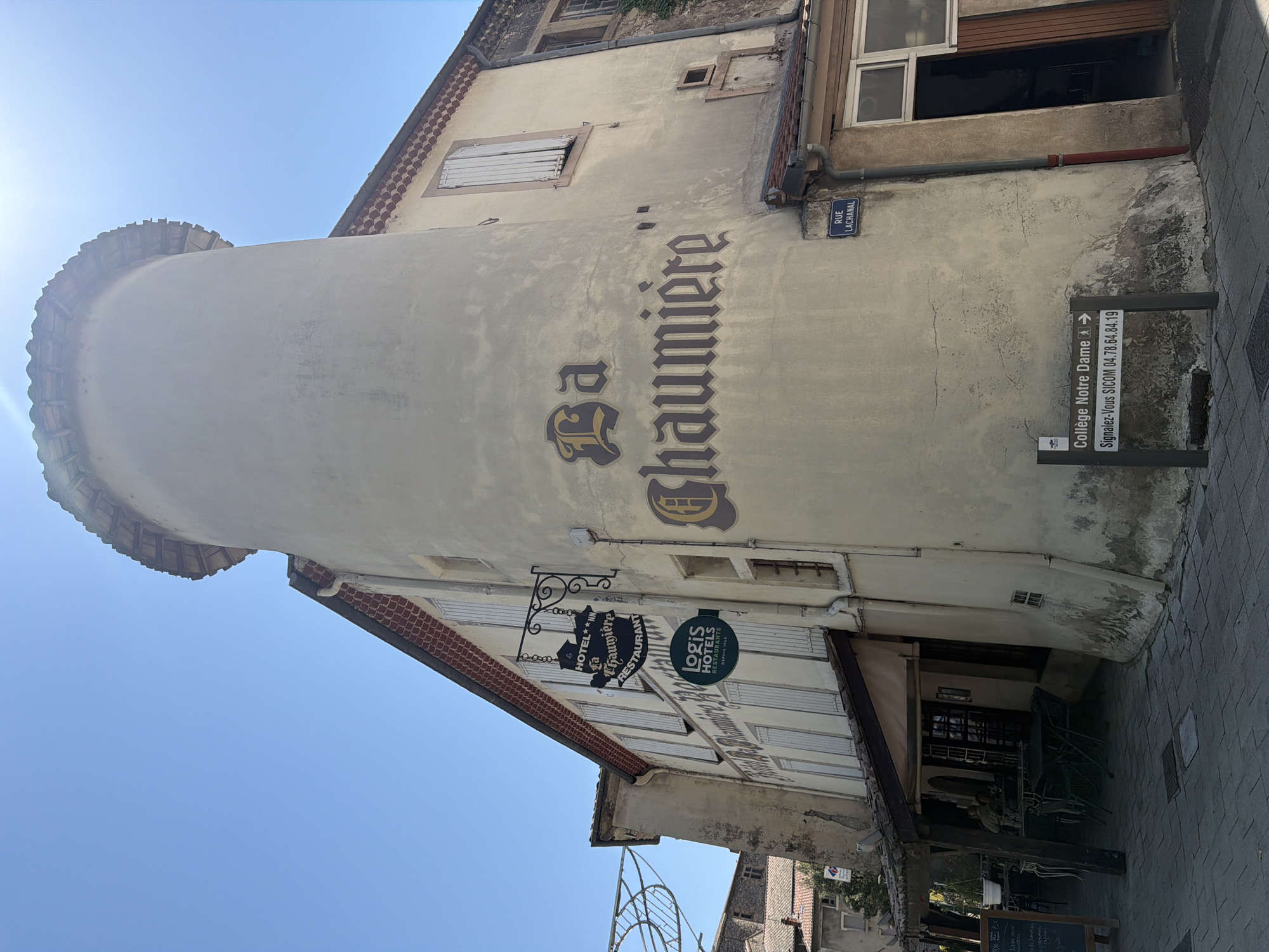 The photograph depicts the exterior of "La Chaumière," a hotel and restaurant located in Tournon-sur-Rhône, France. The building features a distinctive rounded tower with a tiled roof. A wrought-iron sign, bearing the text "HOTEL LA CHAUMIERE RESTAURANT," hangs from the facade. Below this, a circular green sign reads "logis HOTELS RESTAURANTS." Large, ornate lettering on the building's exterior spells out "La Chaumière." A street sign indicating "Rue Lachanal" is visible on the building's corner. Further down, a directional sign points towards "Collège Notre Dame." The scene is set on a paved street, with outdoor seating areas for the restaurant visible under a covered patio. The sky is clear and blue, indicating a sunny day.