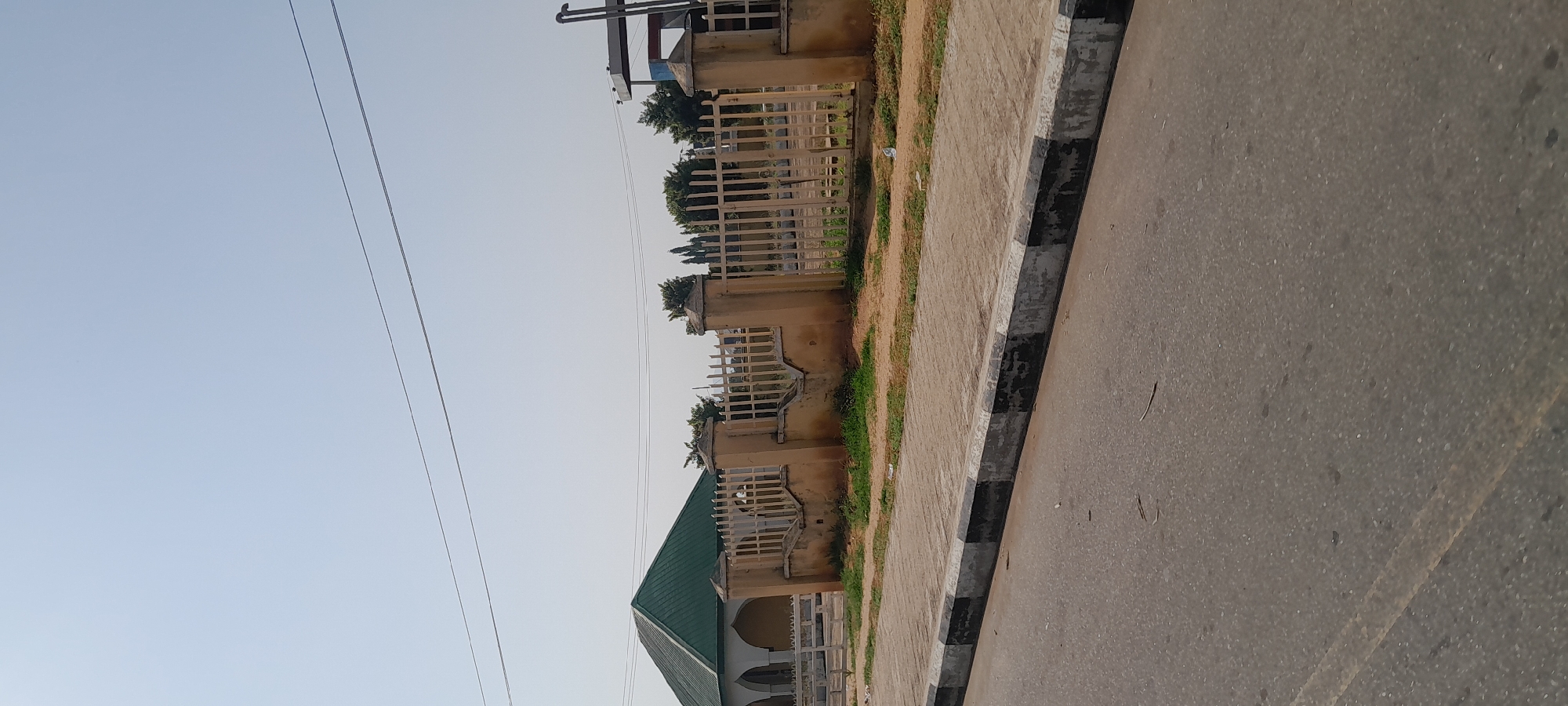 The image shows a street scene in Jalingo, Nigeria. In the background, a building with a green, sloped roof is visible, featuring arched windows and a light-colored facade. A beige metal fence runs along the front of the property, supported by concrete pillars. The fence is partially obscured by vegetation. In the foreground, a paved sidewalk with a black and white striped border separates the street from the fenced property. The street itself is asphalt and appears to be a public road. The sky is clear and light blue, indicating a daytime scene. There are no people or vehicles clearly visible in the frame. The overall impression is of an ordinary urban environment in Nigeria.