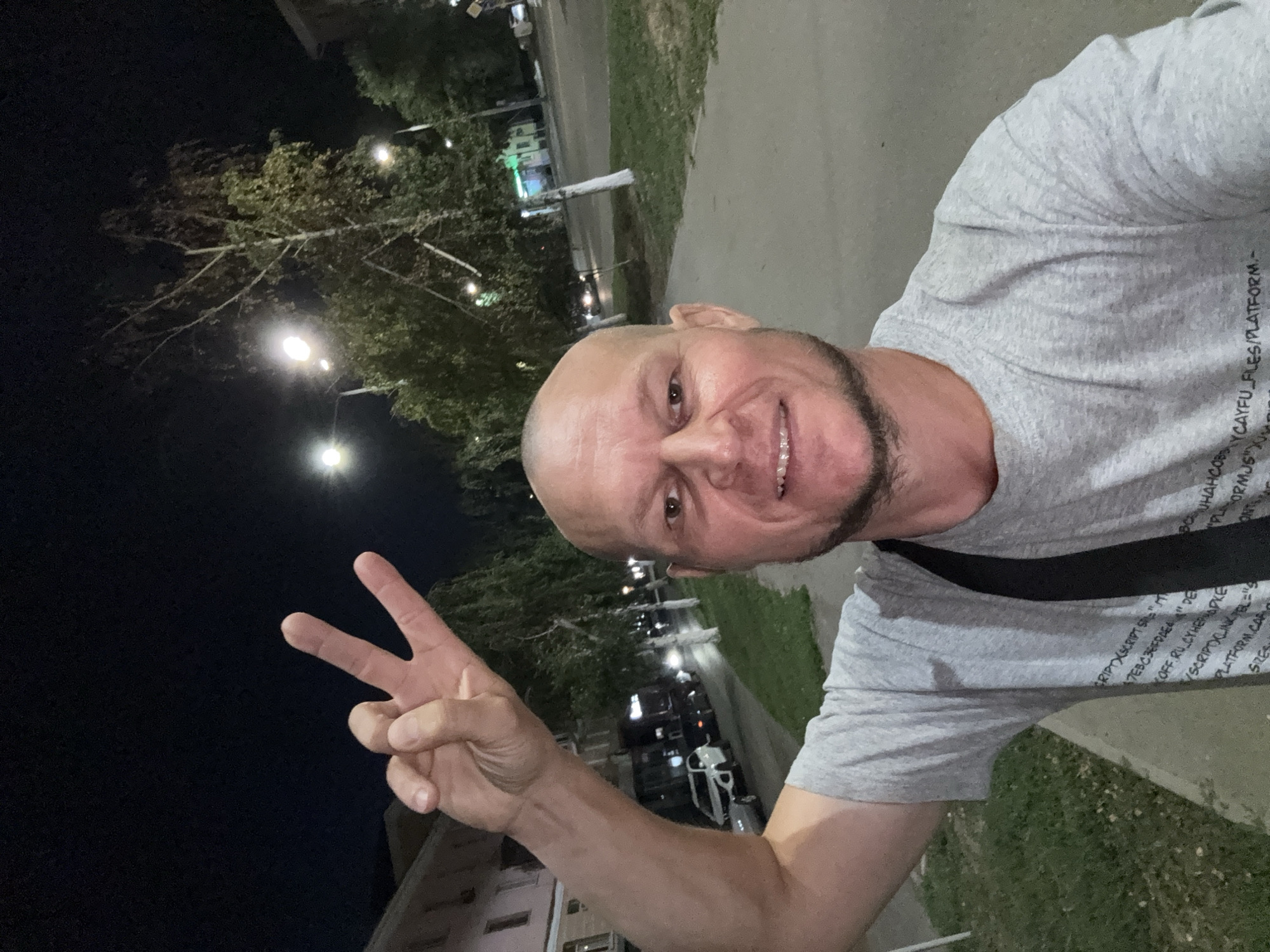 A bald, light-skinned man with a goatee and a slight smile is taking a selfie at night on a sidewalk in Oskemen, Kazakhstan. He is wearing a gray t-shirt with text printed on it, and a black strap is visible across his chest. His right arm is raised, and he is making a peace sign with his hand. The background shows a street with parked cars, streetlights, and trees with white-painted trunks. Buildings are visible on the left side of the street. The scene appears to be a typical urban environment at night.