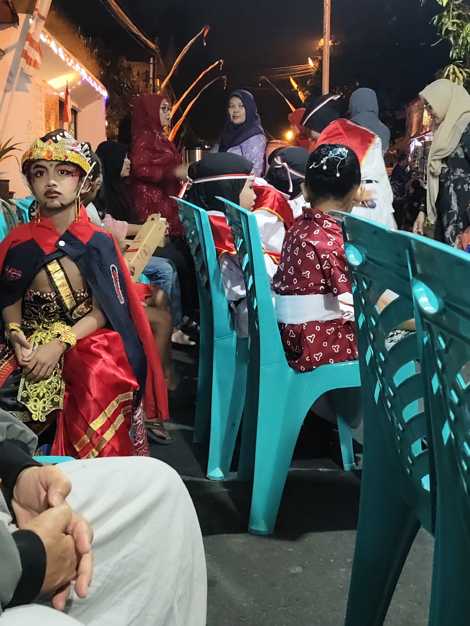 In the city of Magelang, Indonesia, an outdoor nighttime scene is depicted, possibly during a cultural event or festival. The foreground is occupied by a person in traditional Javanese attire, featuring a golden headdress, ornate vest, and red and gold fabric. This individual appears to be a child or young person, with theatrical makeup. They are seated, with hands resting on their lap, positioned behind a bright teal plastic chair.
Further back, several individuals are visible. To the left, a woman in a dark red hijab and glasses observes the scene. Behind her, other people are partially visible, some wearing dark clothing and head coverings. In the center-right, a person wearing a dark patterned outfit and a light-colored hijab is conversing with another individual whose face is obscured. Another person, likely a child, is seated in a red and white patterned garment with a white sash, facing away from the camera.
The background shows a building adorned with Indonesian flags and lights, suggesting a celebratory atmosphere. The ground appears to be asphalt or pavement. The lighting is dim, with artificial light sources illuminating the area. The overall impression is of a community gathering in an urban or semi-urban setting in Magelang.