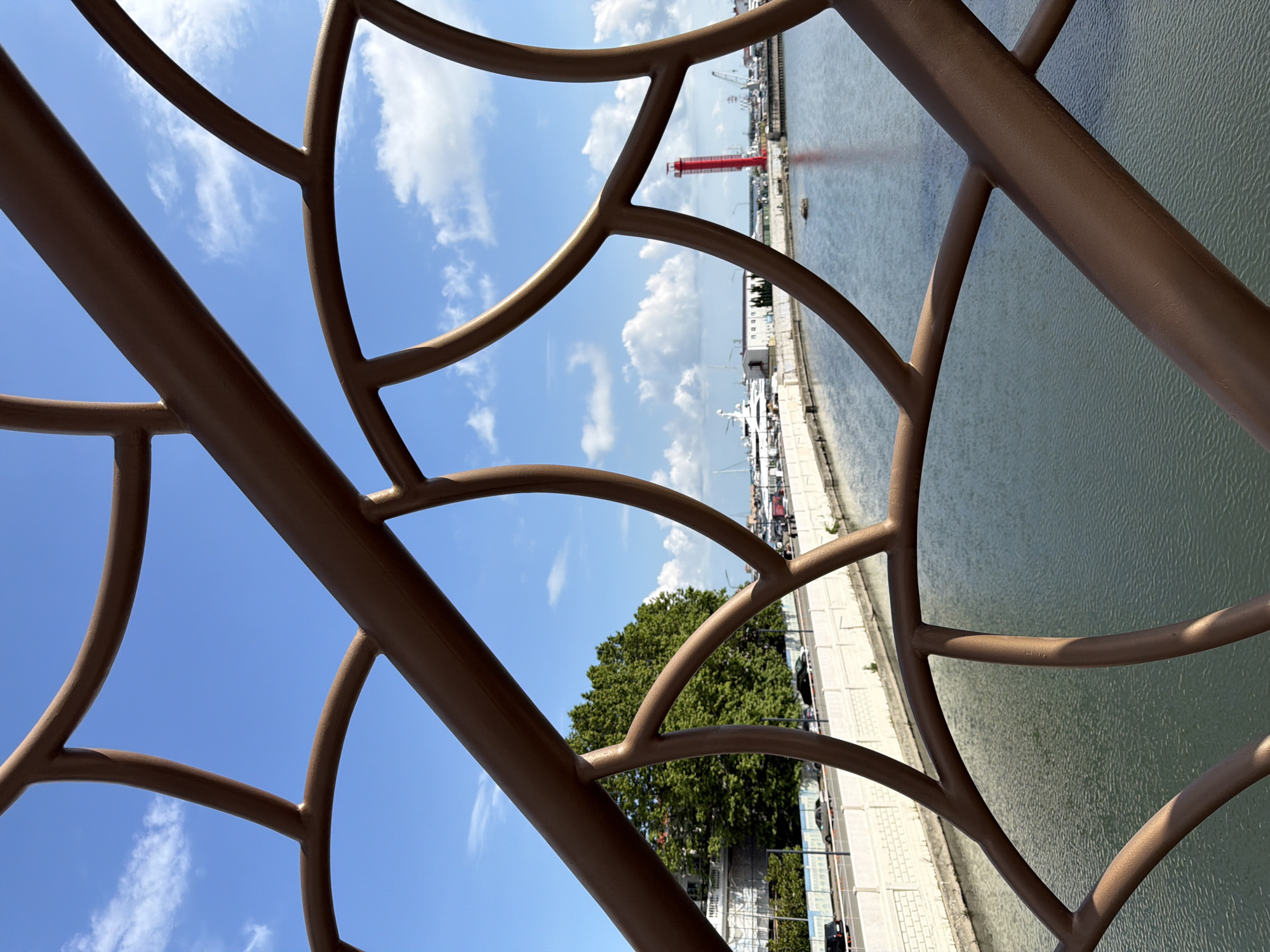 The image captures a view of the Sochi Sea Port in Russia, framed by the decorative, bronzed metalwork of a bridge railing. The railing features a repeating, organic pattern resembling branches or veins. Through the gaps in the railing, a bright blue sky with scattered white clouds is visible.
Below the railing, a calm, blue body of water, likely the Black Sea, reflects the sky. Along the waterfront, a white embankment wall runs parallel to the water, with a road above it. Several cars are seen driving on this road. Further in the background, lush green trees and white buildings line the shore.
Dominating the port scene are several large white yachts moored in the harbor. To the right of the yachts, a distinctive red lighthouse stands tall, a prominent landmark of the port. Other industrial structures and cranes are visible in the distance, indicating the active nature of the port. The overall scene suggests a sunny day with moderate activity at the port.